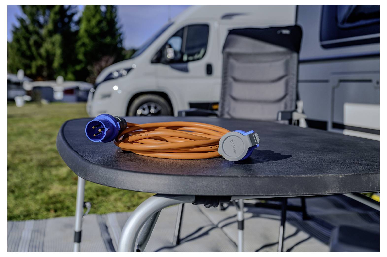 Oranges Stromkabel mit blauen Steckern, aufgewickelt auf einem Tisch neben einem Wohnmobil im Freien.