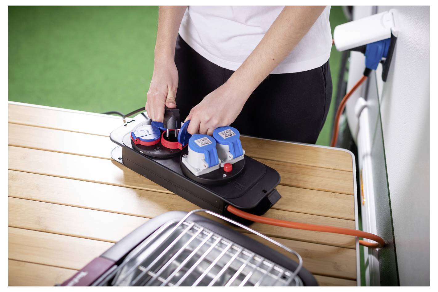 Person, die eine Steckerleiste mit blauen Steckern verwendet, die in einen schwarzen Verteilerstecker auf einem Tisch gesteckt ist, mit angeschlossenem orangefarbenem Stromkabel.