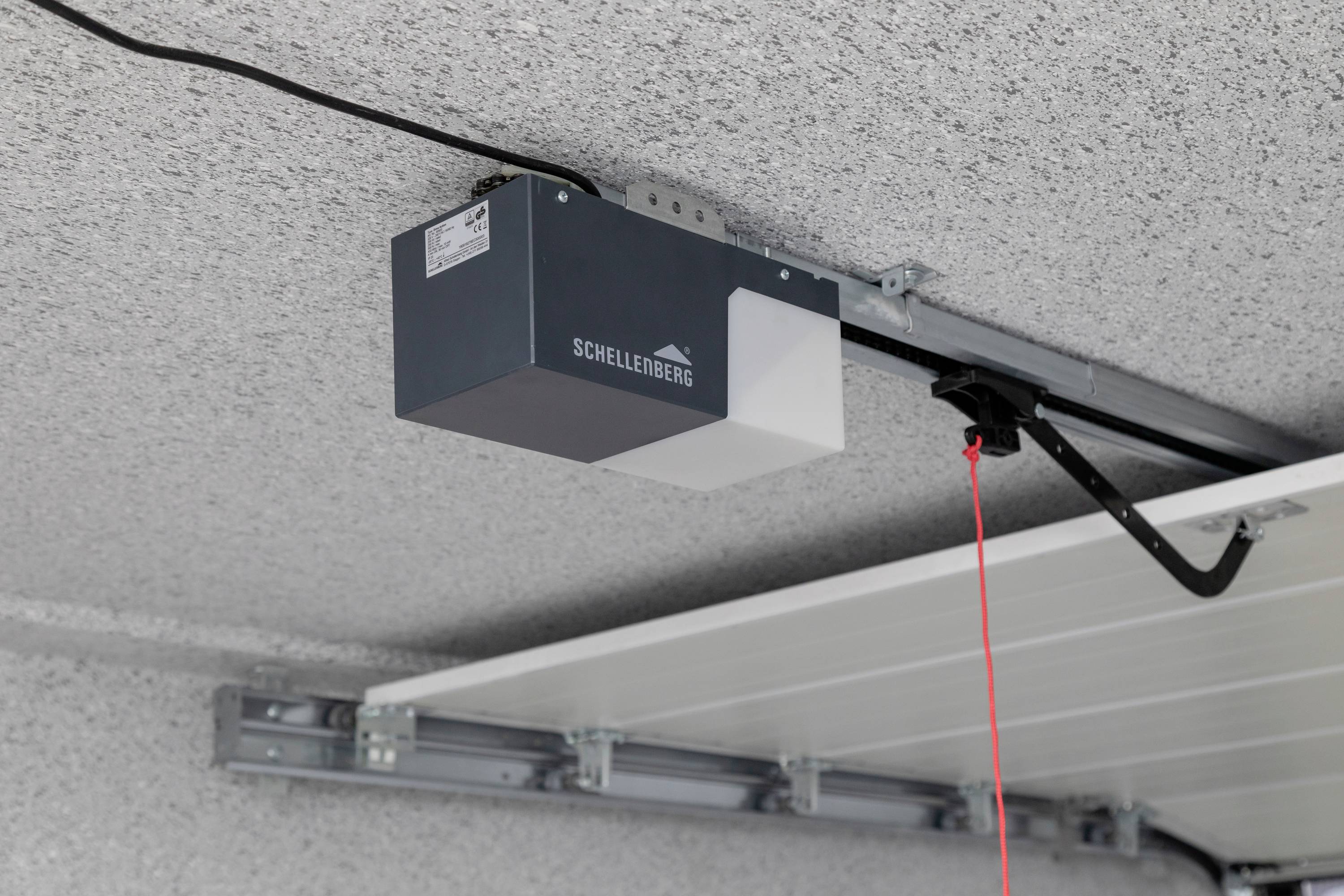 'Elektrischer Garagentorantrieb von Schellenberg an einer Decke montiert, ein rotes Zugseil hängt davon herunter.'