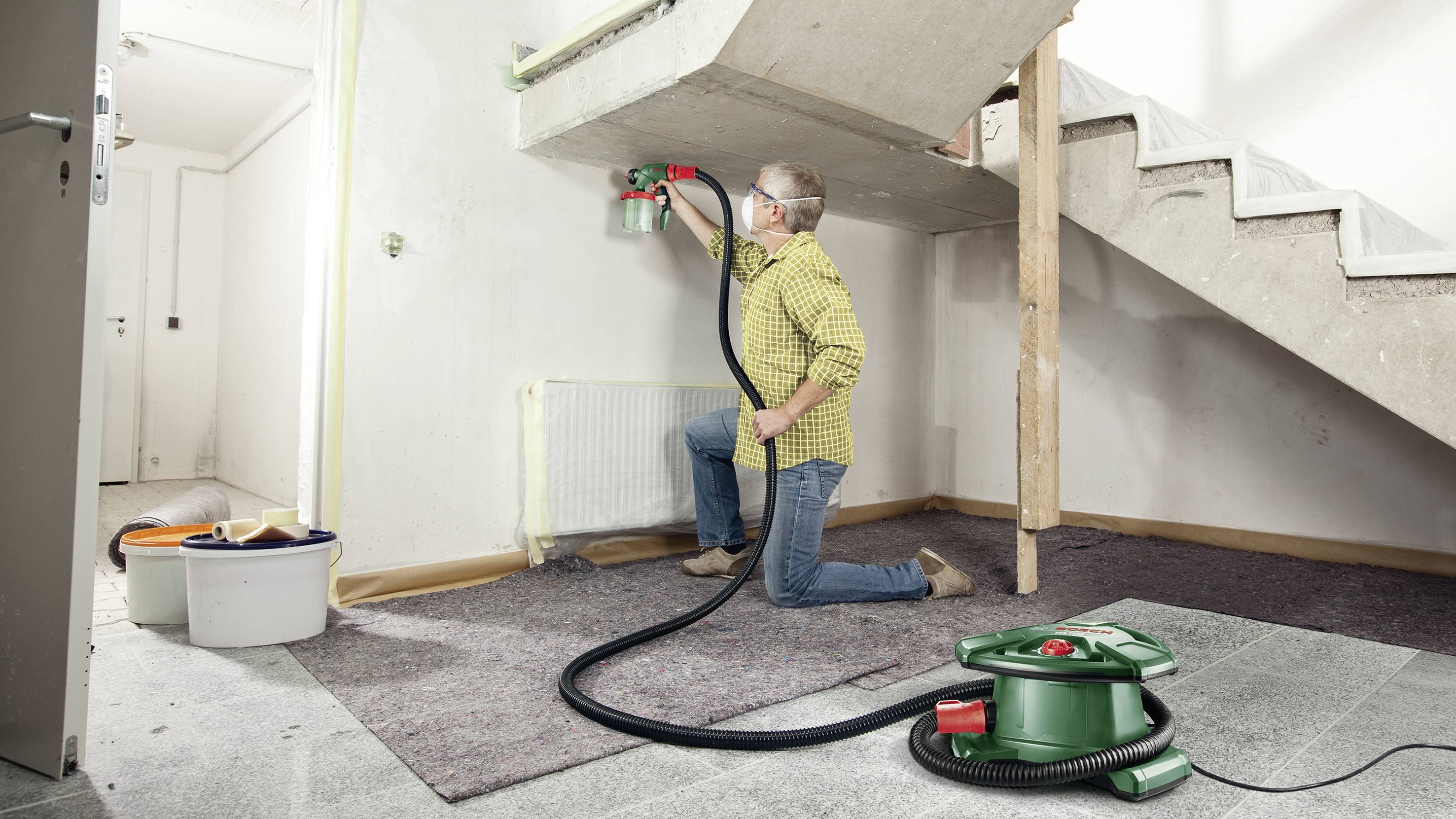 Ein Mann in Schutzbrille sprüht Farbe auf eine Wand unter einer Treppe. Ein Farbkompressor steht am Boden. Renovierungsszene in einem Zimmer.