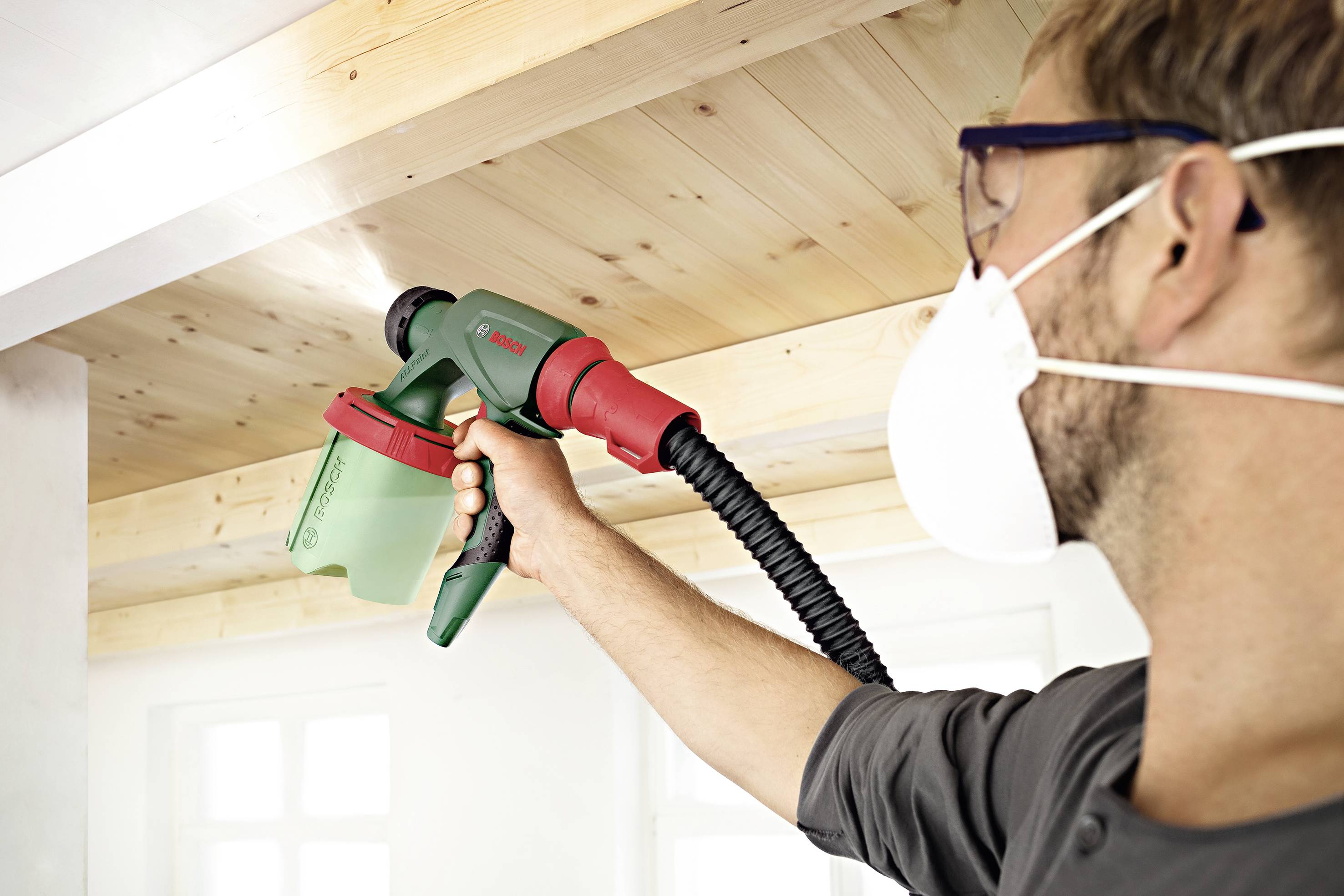 Eine Person mit Schutzbrille und Maske sprüht mit einem grünen und roten Farbsprüher Farbe an eine Holzdecke in einem hellen Raum.