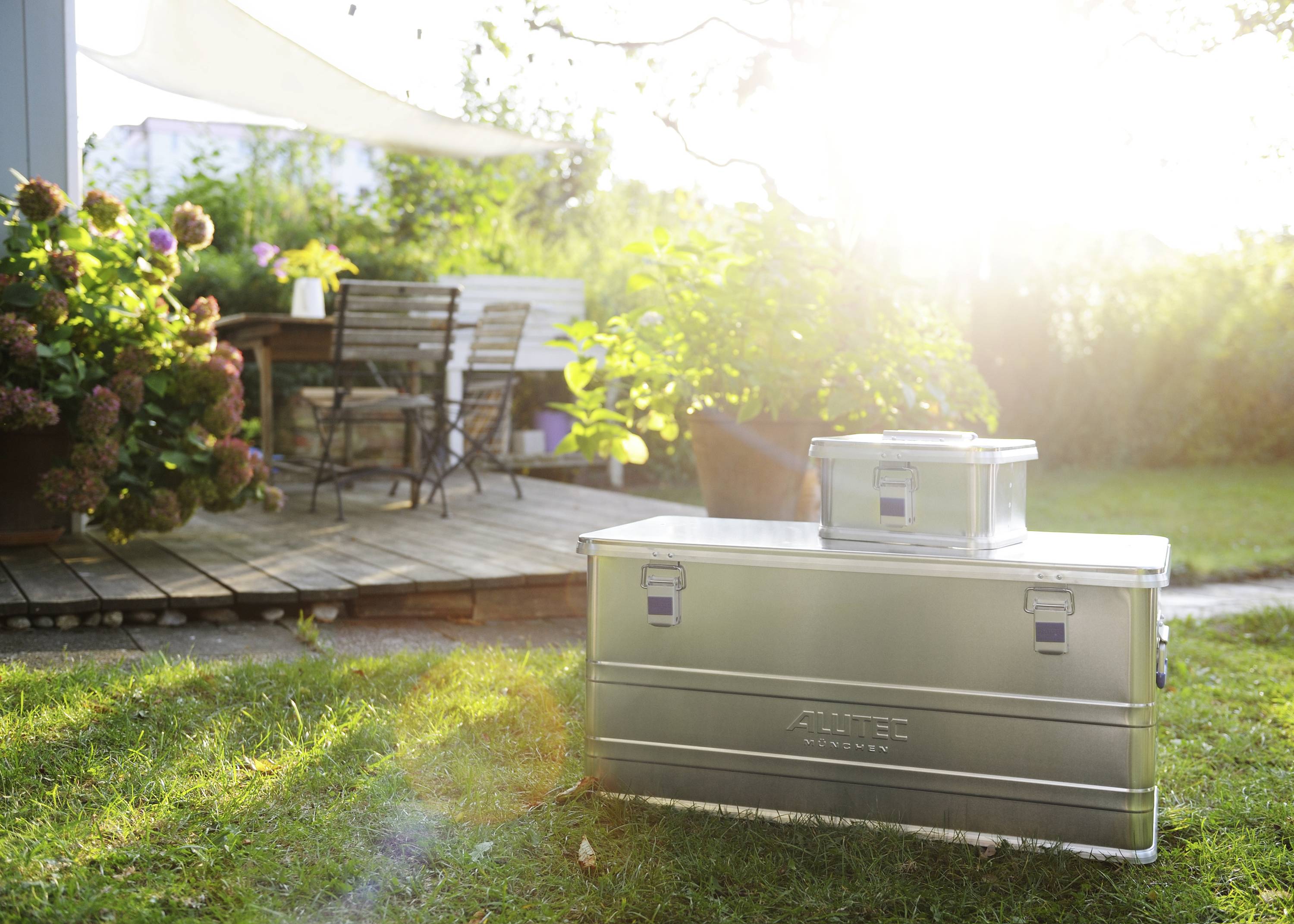 Ein sonniger Garten mit blühenden Pflanzen und Holzmöbeln auf einer Terrasse. Zwei Metallkisten stehen auf dem Rasen im Vordergrund.