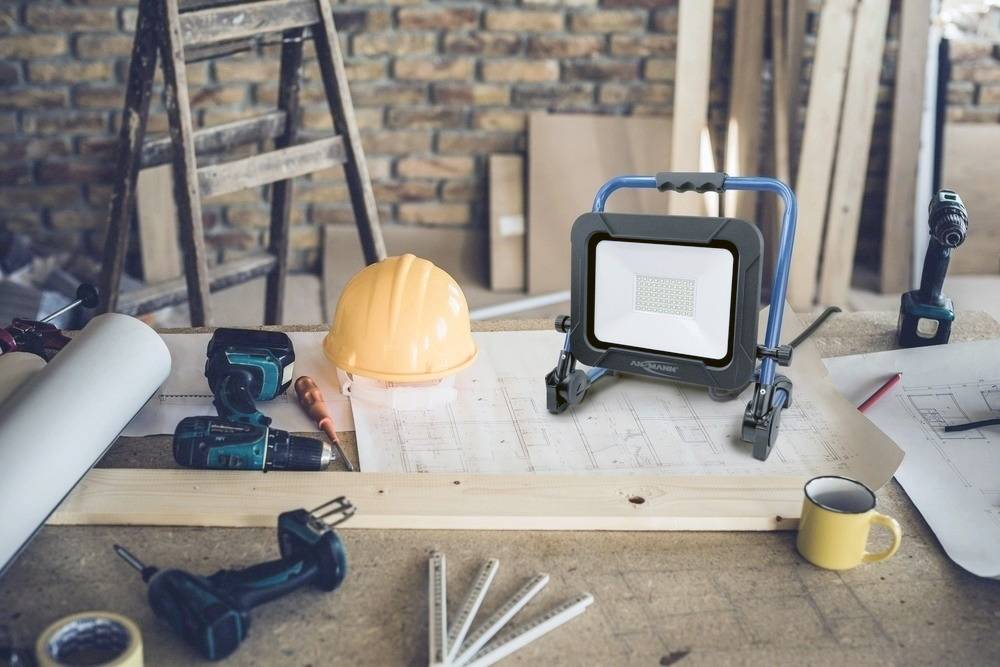 Werkbank mit Bauplänen, Schutzhelm, Taschenlampe, Bohrer und Kaffeetasse. Im Hintergrund eine Ziegelwand und eine Leiter.