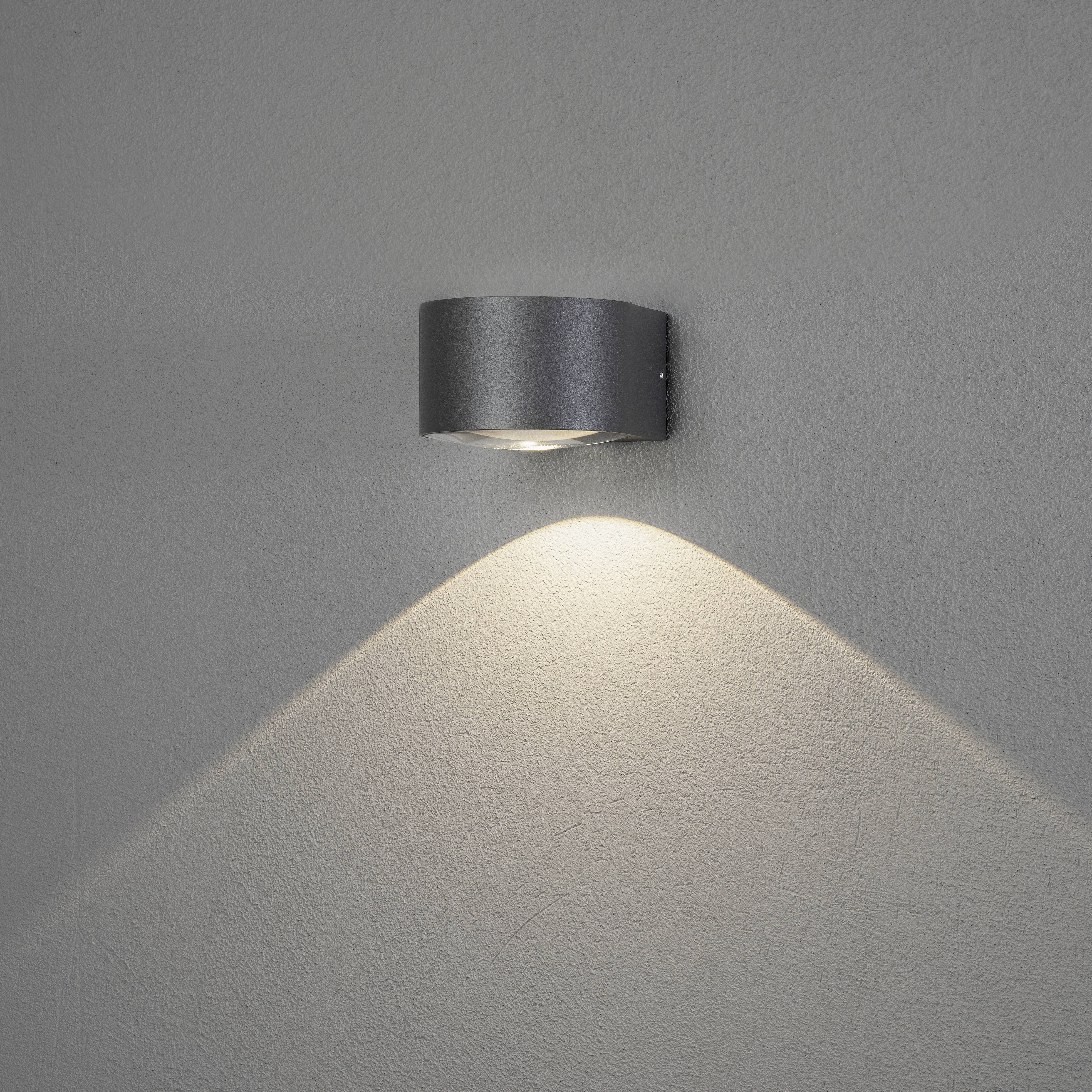 Eine silberne Wandleuchte strahlt warmes Licht nach unten auf eine graue Wand, wodurch ein gleichmäßiger Lichteffekt entsteht.