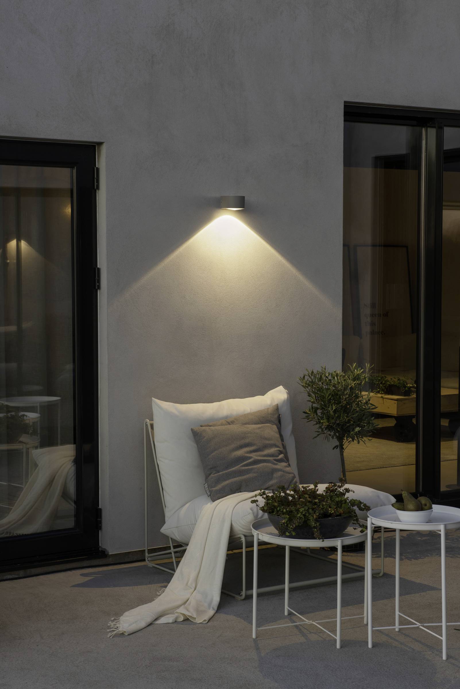 Ein gemütlicher Außenbereich mit Sessel, Decke und Kissen unter einer leuchtenden Wandlampe; Glasfenster im Hintergrund.