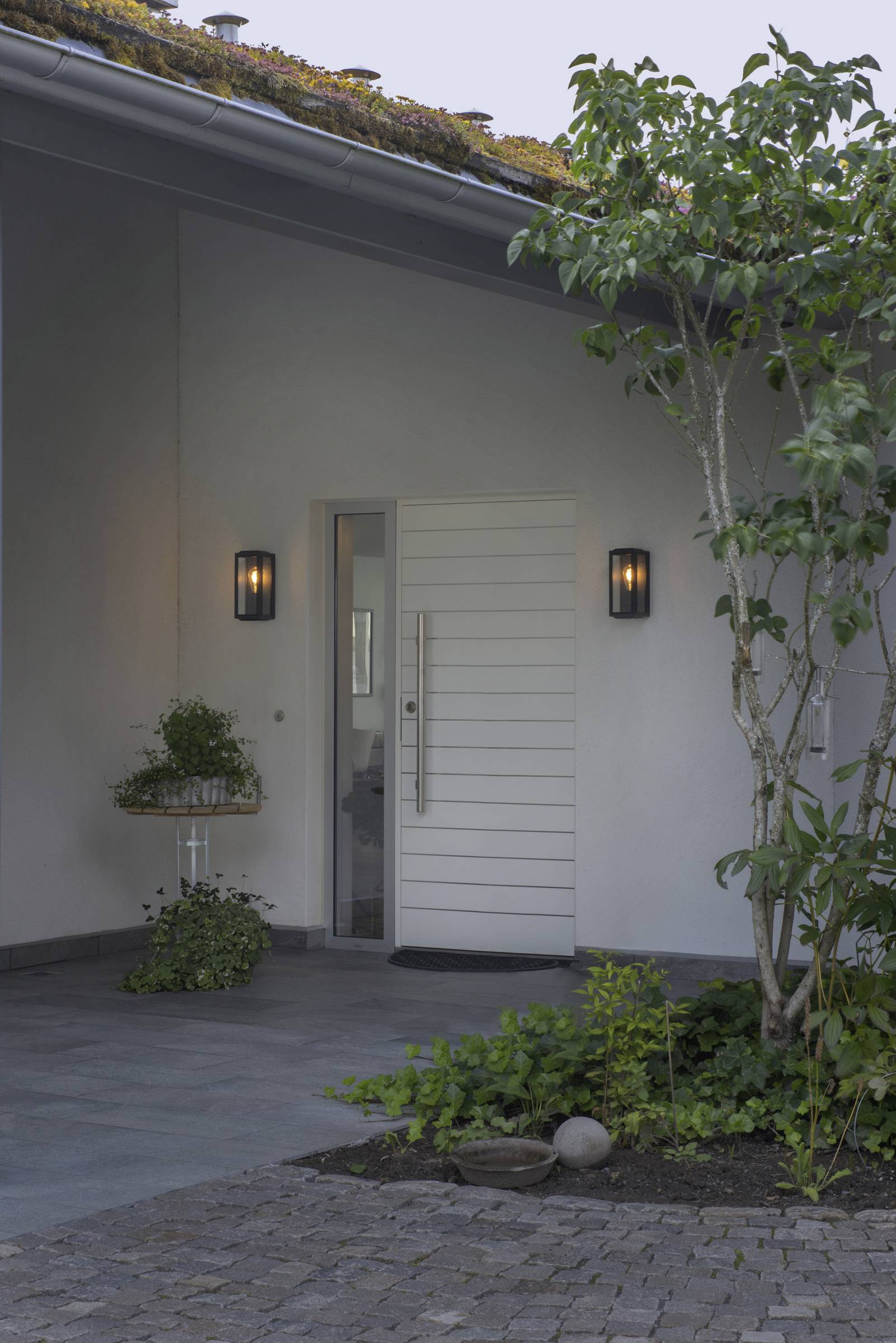 Moderner Hauseingang mit weißer Tür, flankiert von zwei Wandlampen. Links Pflanzen auf einem kleinen Tisch. Rechter Vordergrund: Baum und Pflanzenbeet.