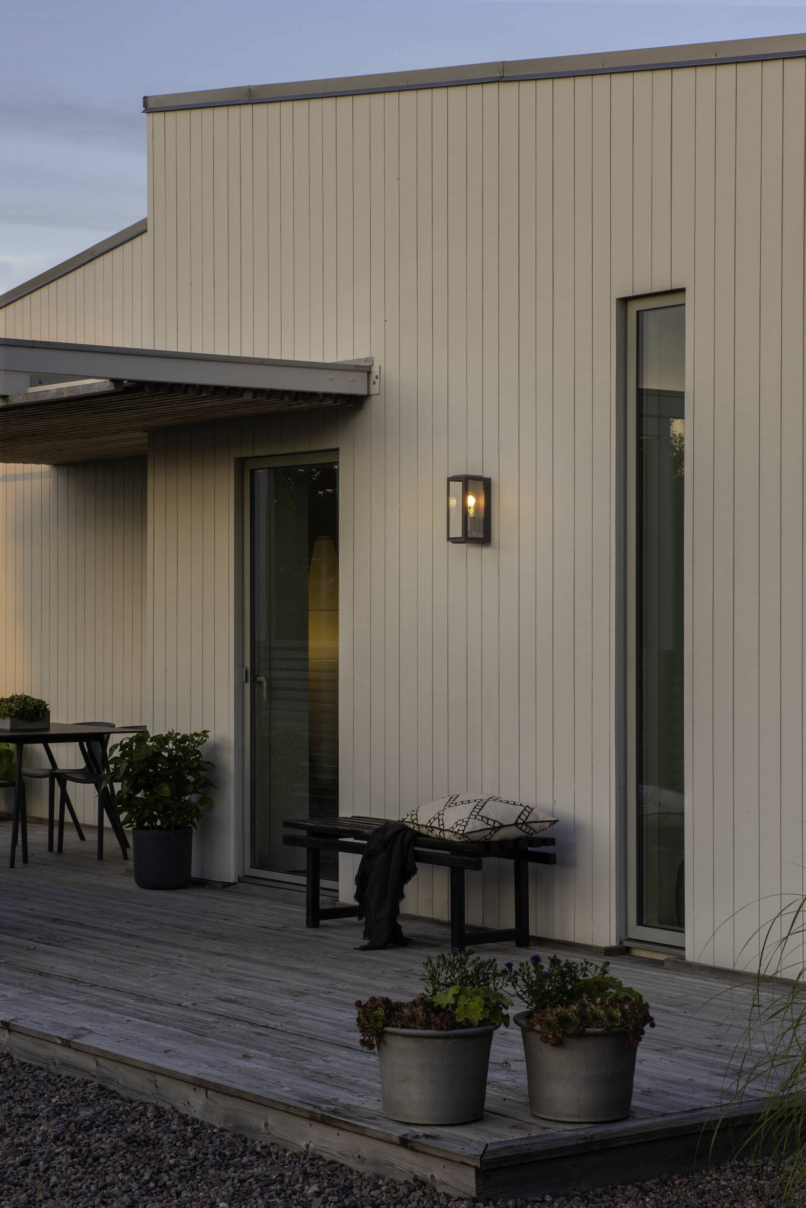 Modernes Haus mit Holzverkleidung bei Dämmerung. Eine Terrasse mit Pflanzen, zwei Stühle und ein Tisch sind zu sehen.