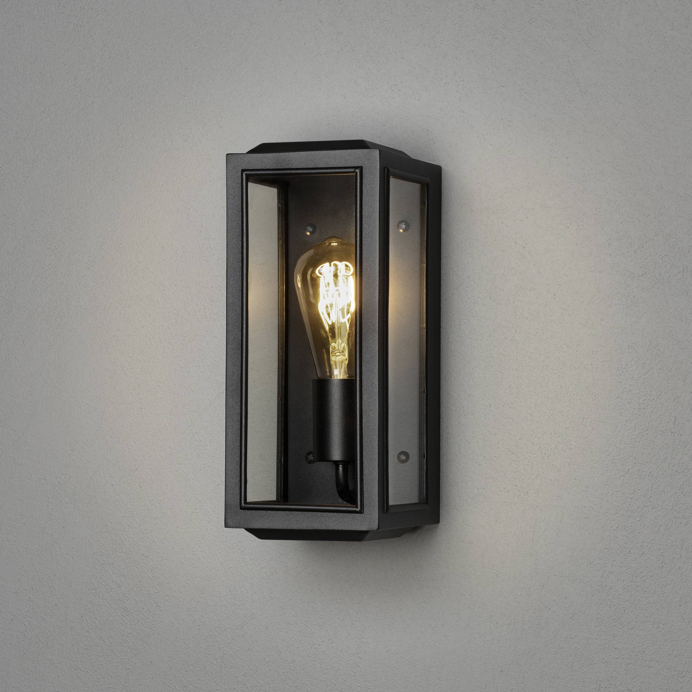 Wandleuchte mit Glasbox-Design, sichtbarem Glühbirnenlicht und schwarzem Metallrahmen an grauer Wand.