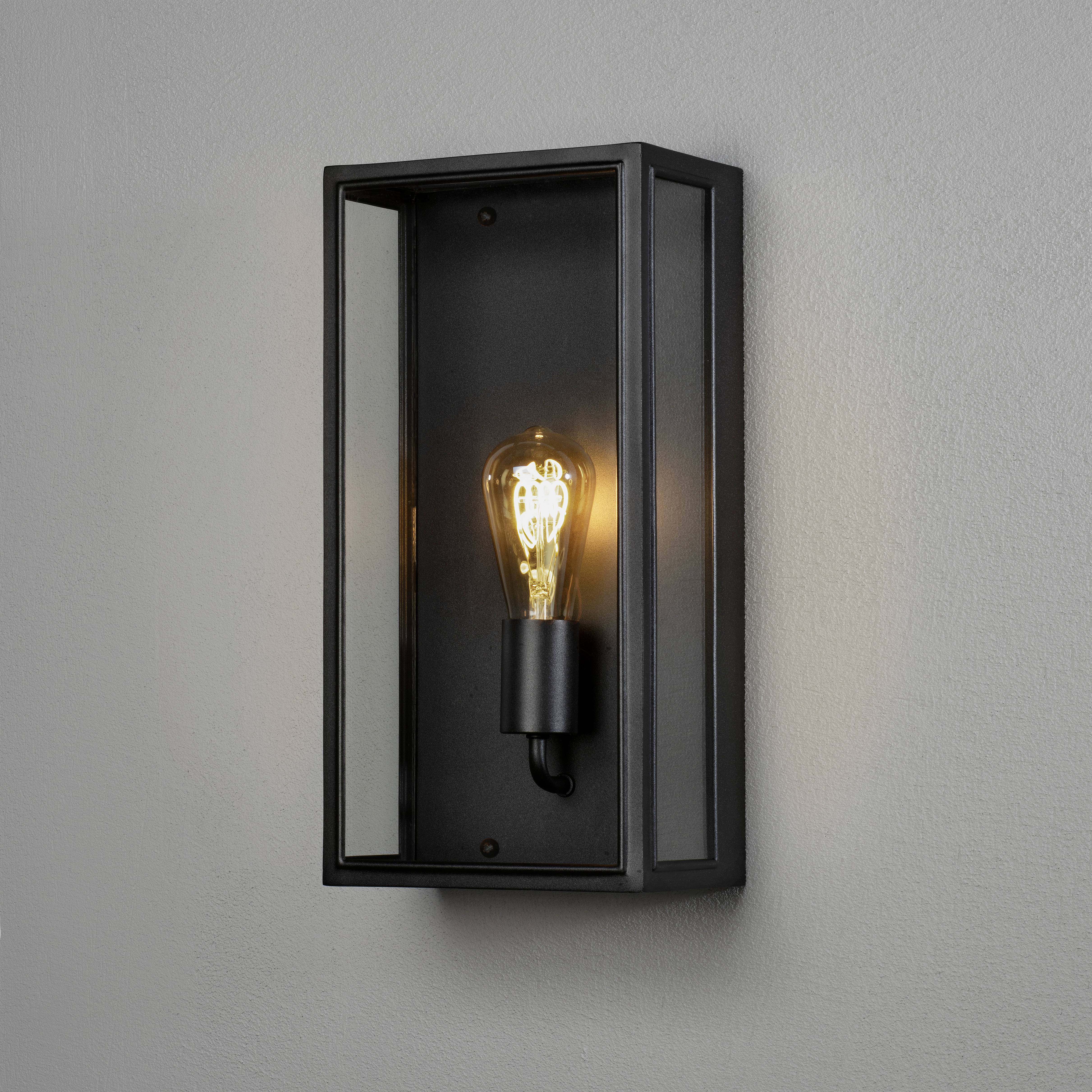 Wandlampe mit schwarzem Rahmen und freiliegender Glühbirne, leuchtet warmes Licht aus; modernes Design.