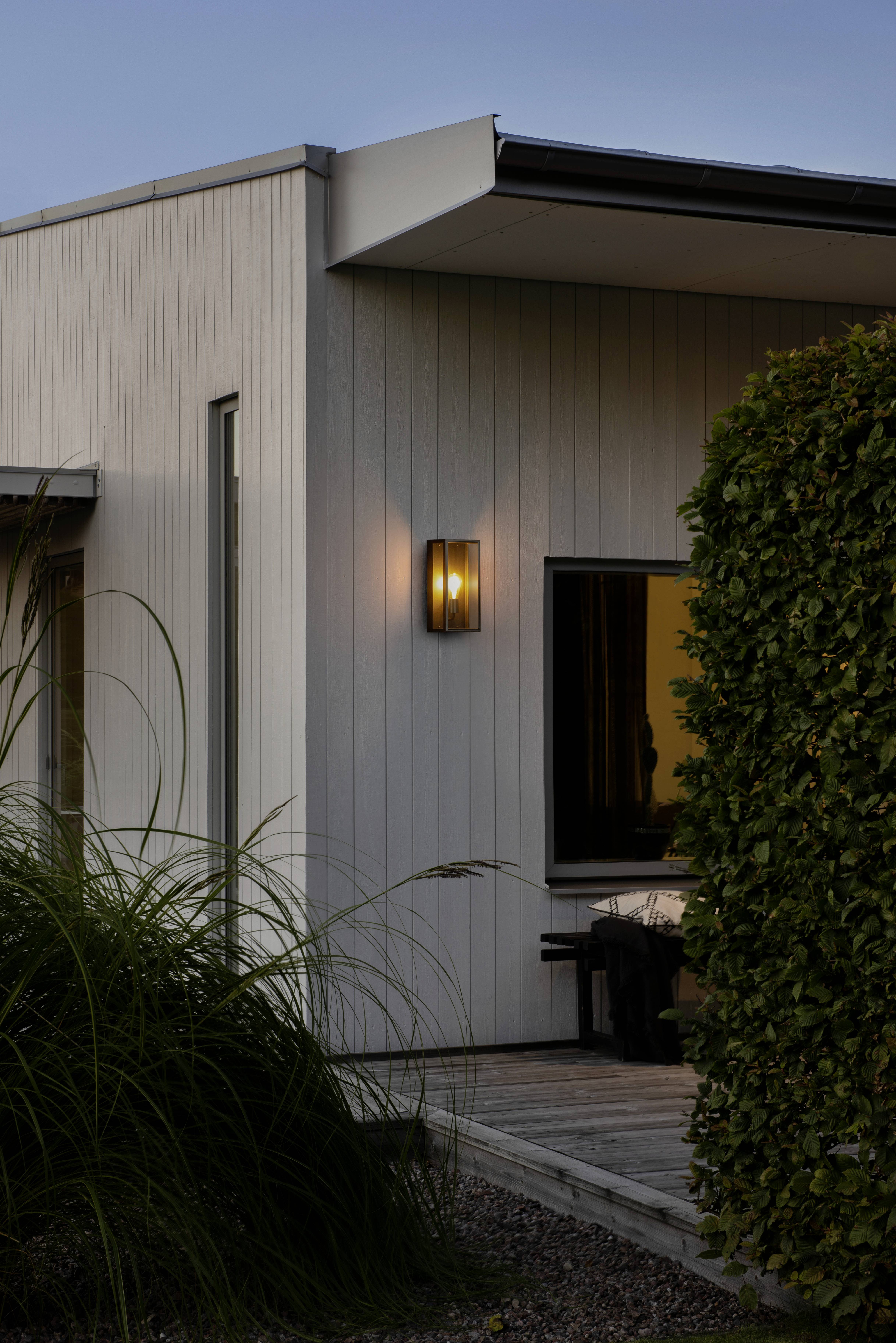 Modernes, zweistöckiges Gebäude mit beleuchteter Außenlampe und großen Fenstern, umgeben von gepflegten Pflanzen im Vordergrund.