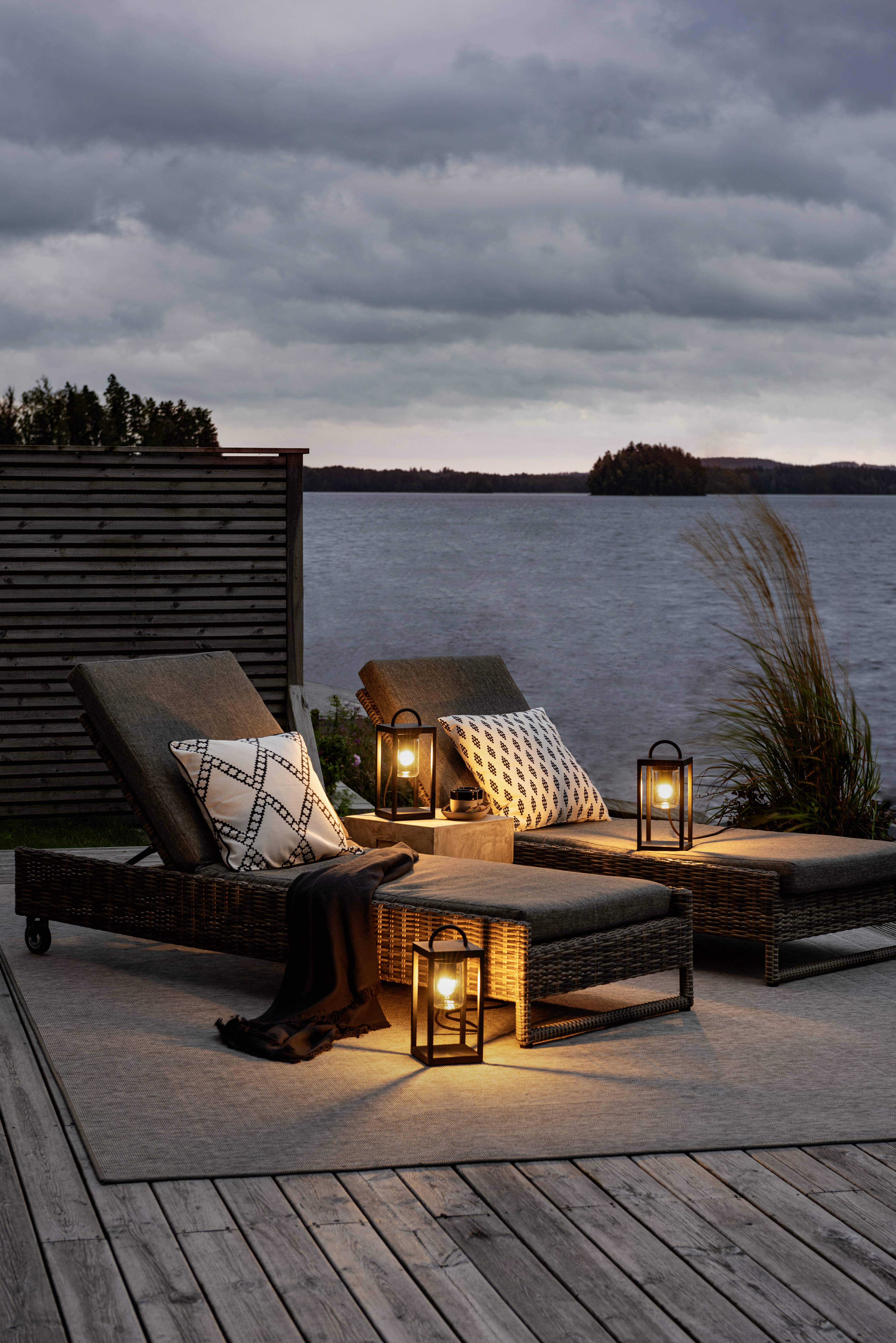 Zwei Liegestühle mit Kissen und Decken auf einer Holzterrasse am See. Laternen leuchten, Wolken am Himmel, Abendstimmung.