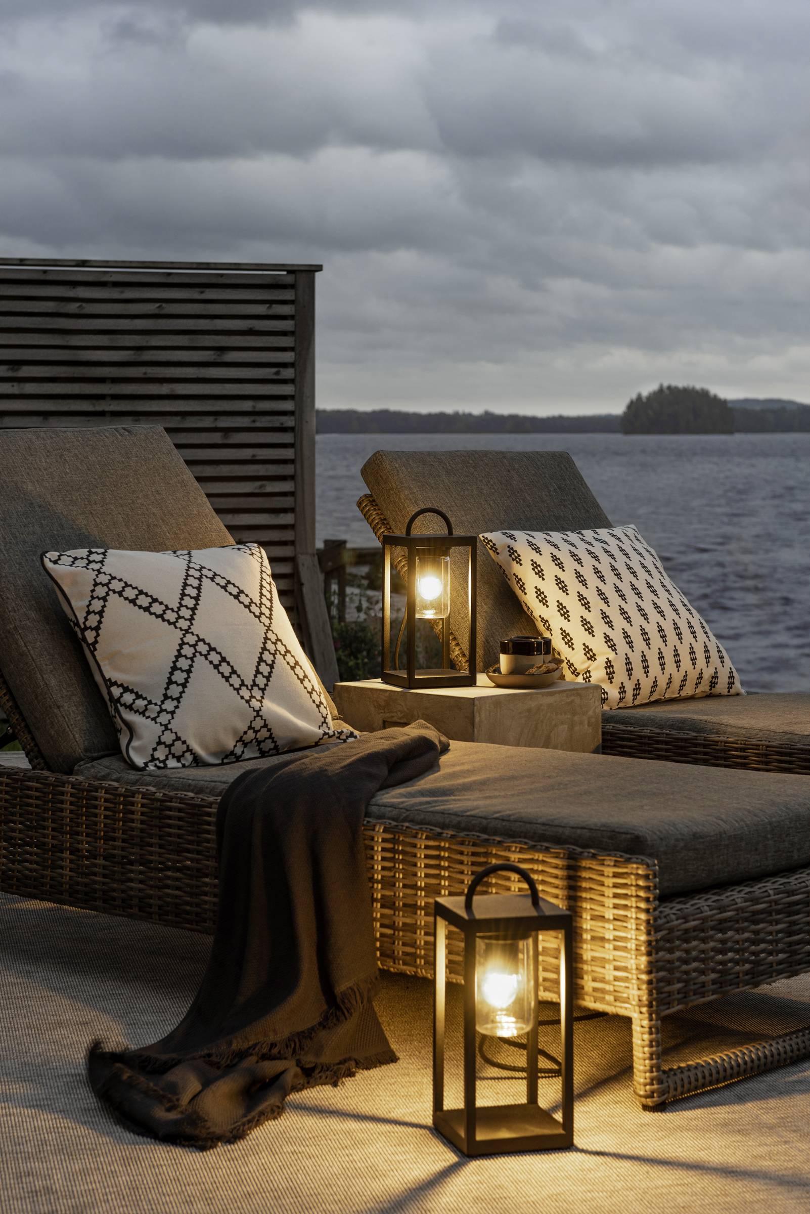 Zwei Liegestühle mit Kissen und Decke stehen auf einer Terrasse am Wasser. Laternen spenden warmes Licht in der Abenddämmerung.