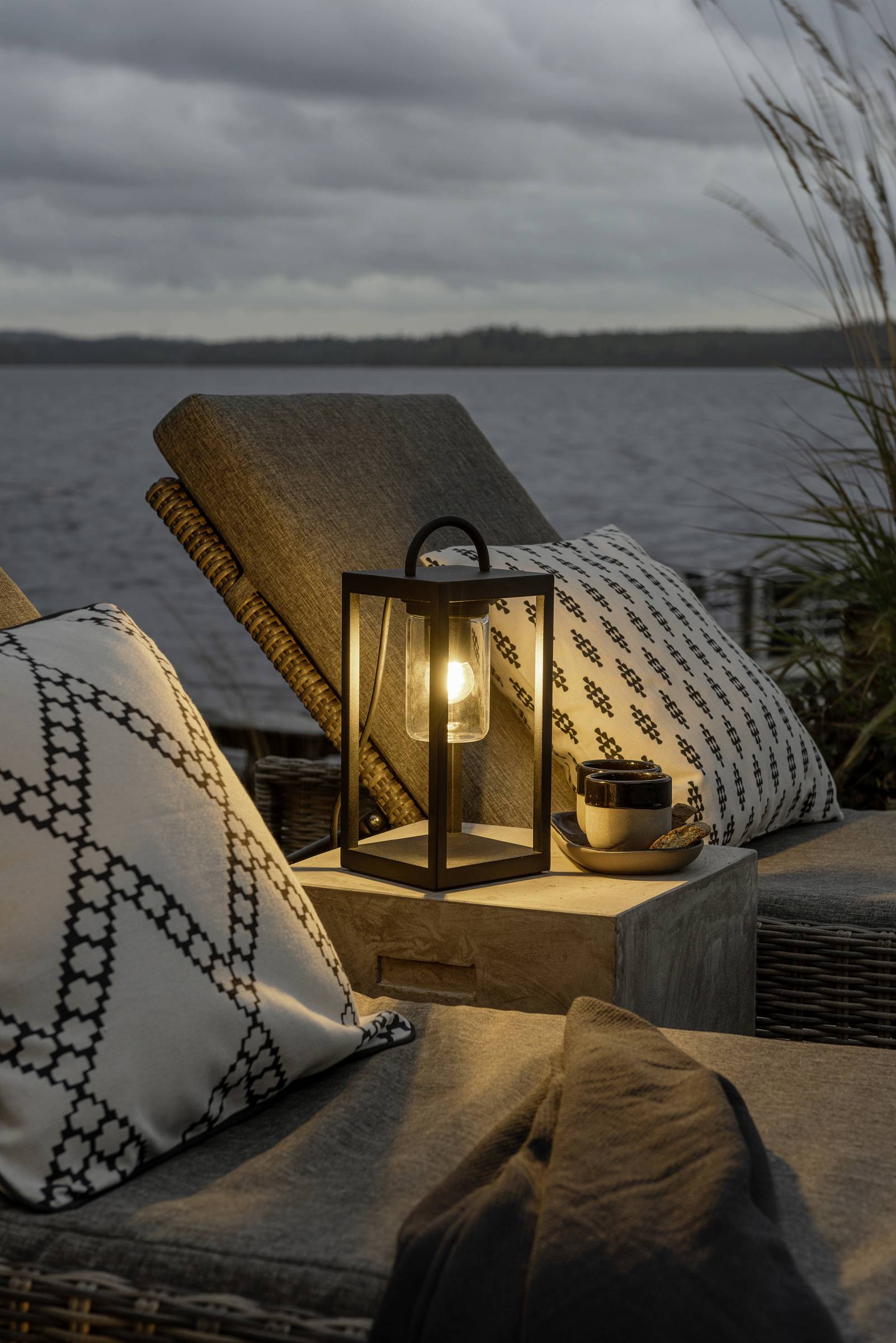 Gemütliche Terrasse mit Rattanmöbeln, dekorativen Kissen, einer leuchtenden Laterne und Tasse Kaffee, im Hintergrund ein See bei Abenddämmerung.
