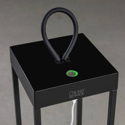 Eine schwarze Tischleuchte mit rechteckiger Basis und geschwungenem Bügelgriff. Ein grüner Knopf mit einem Kabel ist sichtbar.