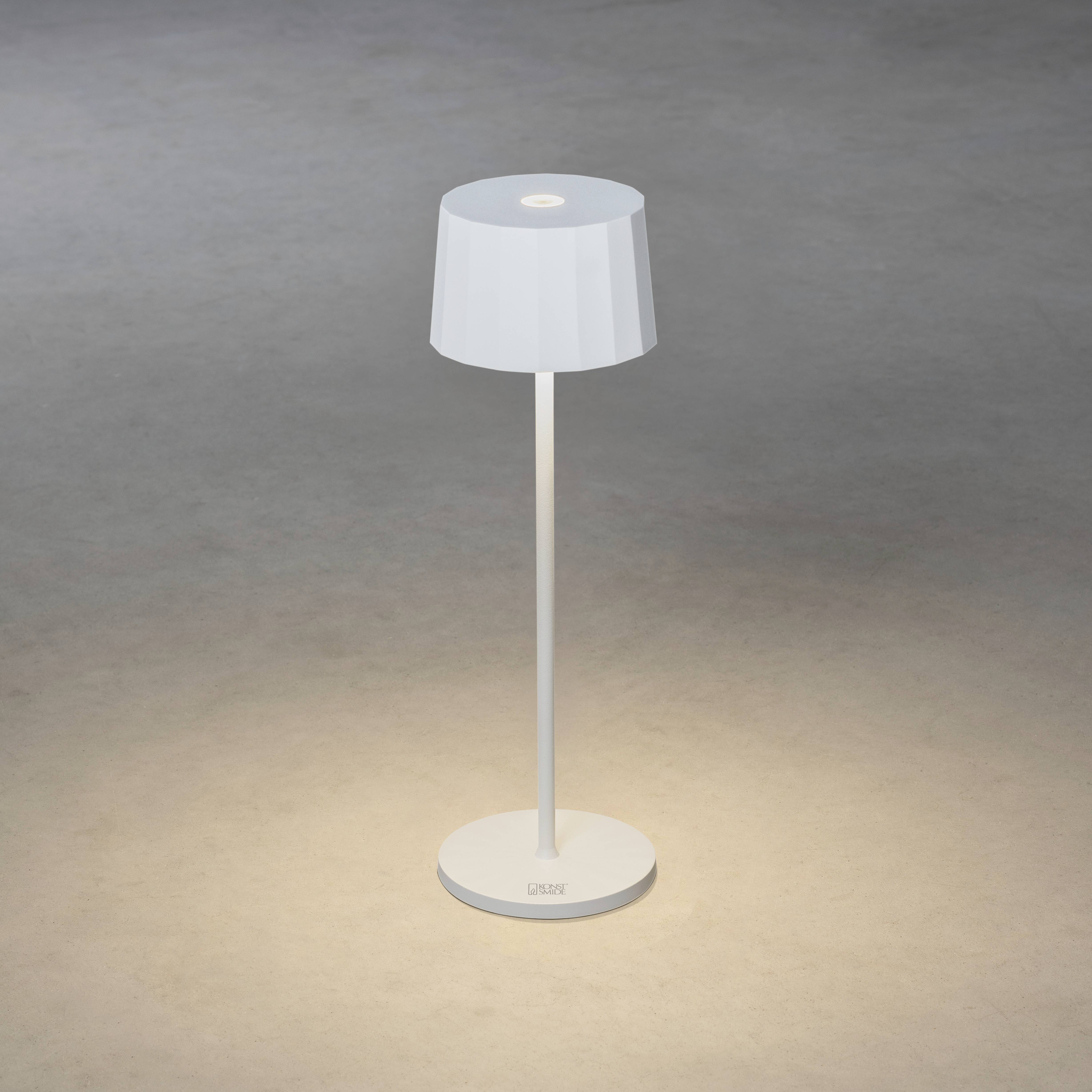 Eine moderne, weiße Tischlampe mit zylindrischem Schirm steht auf einem grau strukturierten Boden, verbreitet weiches Licht.