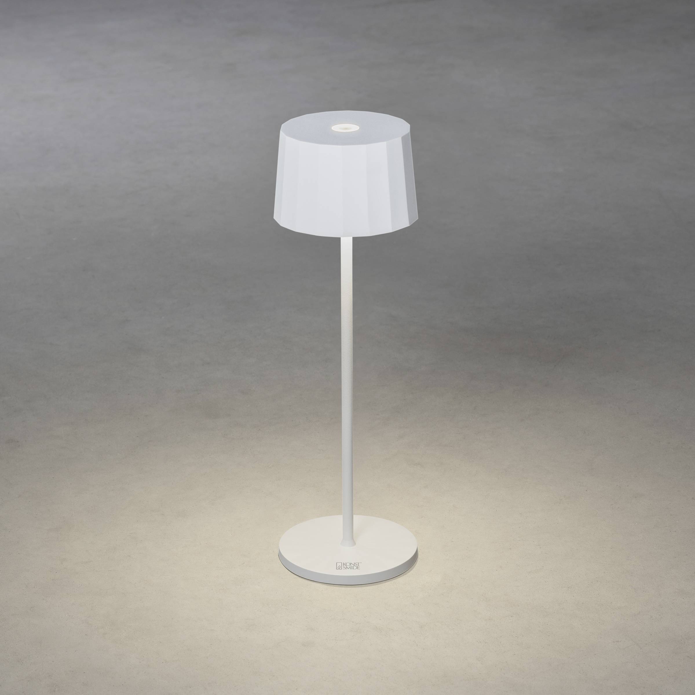 Eine moderne weiße Tischlampe steht auf einem grauen Boden. Sie hat einen zylindrischen Lampenschirm und leuchtet nach unten.