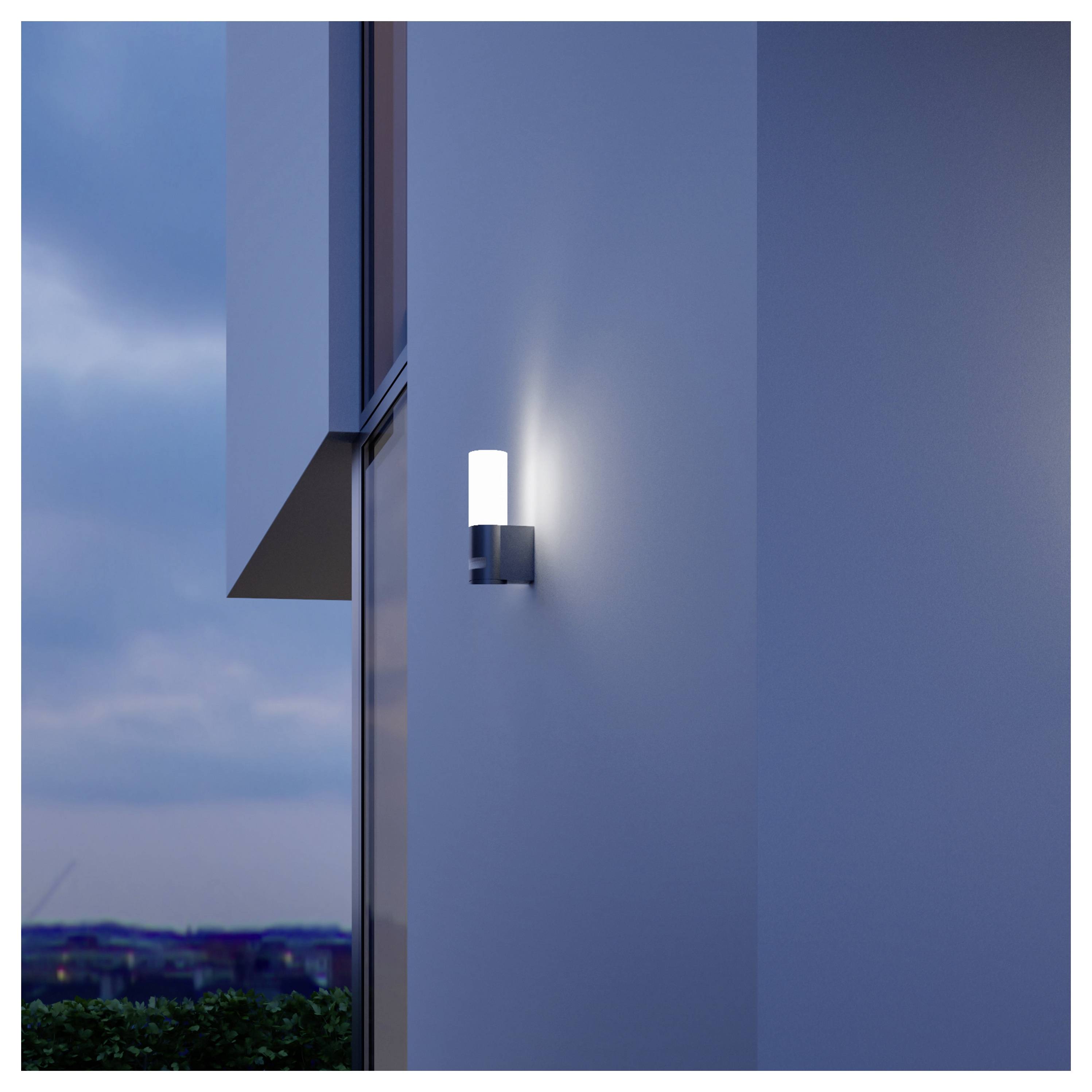Modernes Außenwandlicht, das an einem weißen Gebäude neben einem Fenster in der Dämmerung leuchtet.