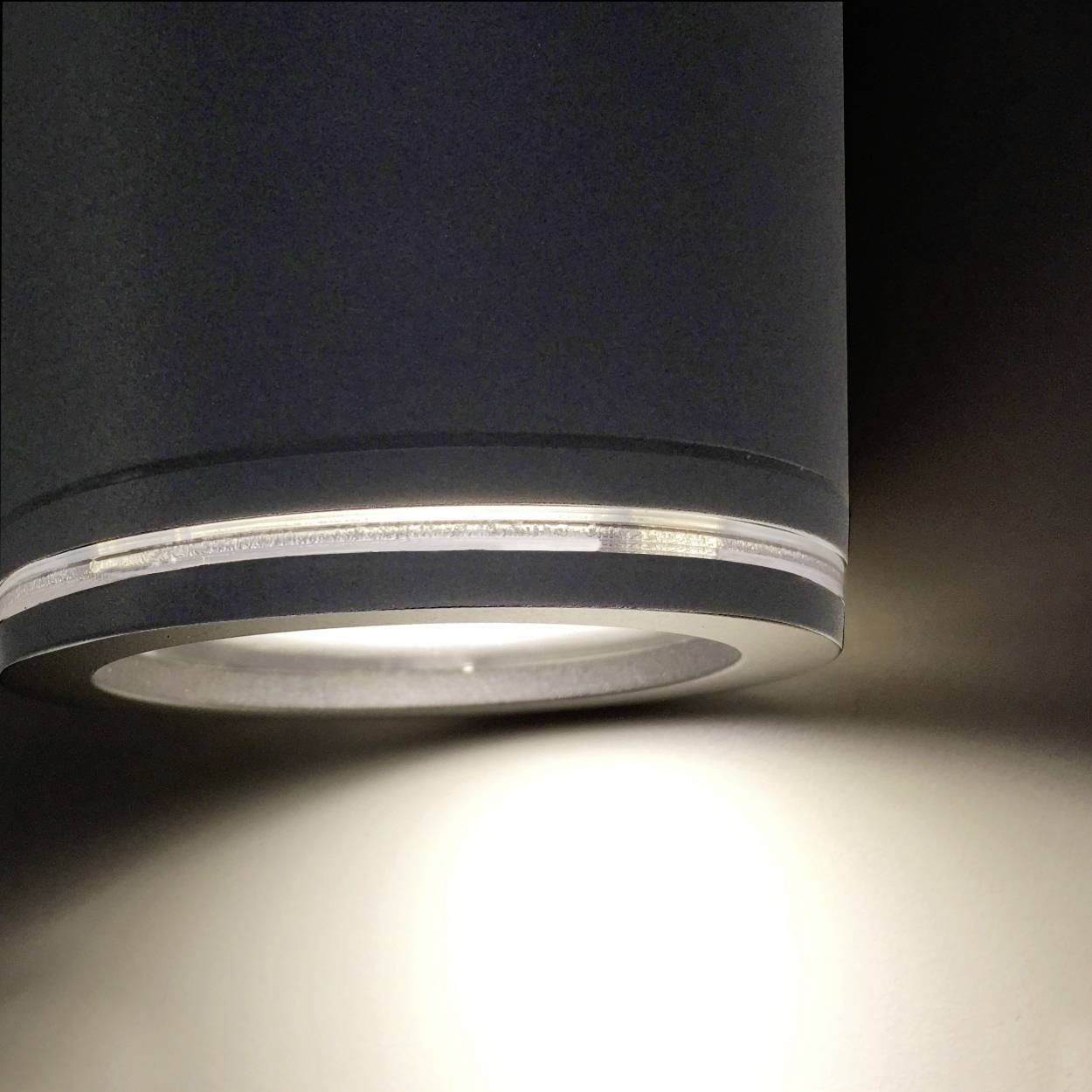 Eine moderne Deckenleuchte mit rundem, grauem Gehäuse strahlt warmes Licht auf eine Wand und erzeugt einen sanften Lichtschein.