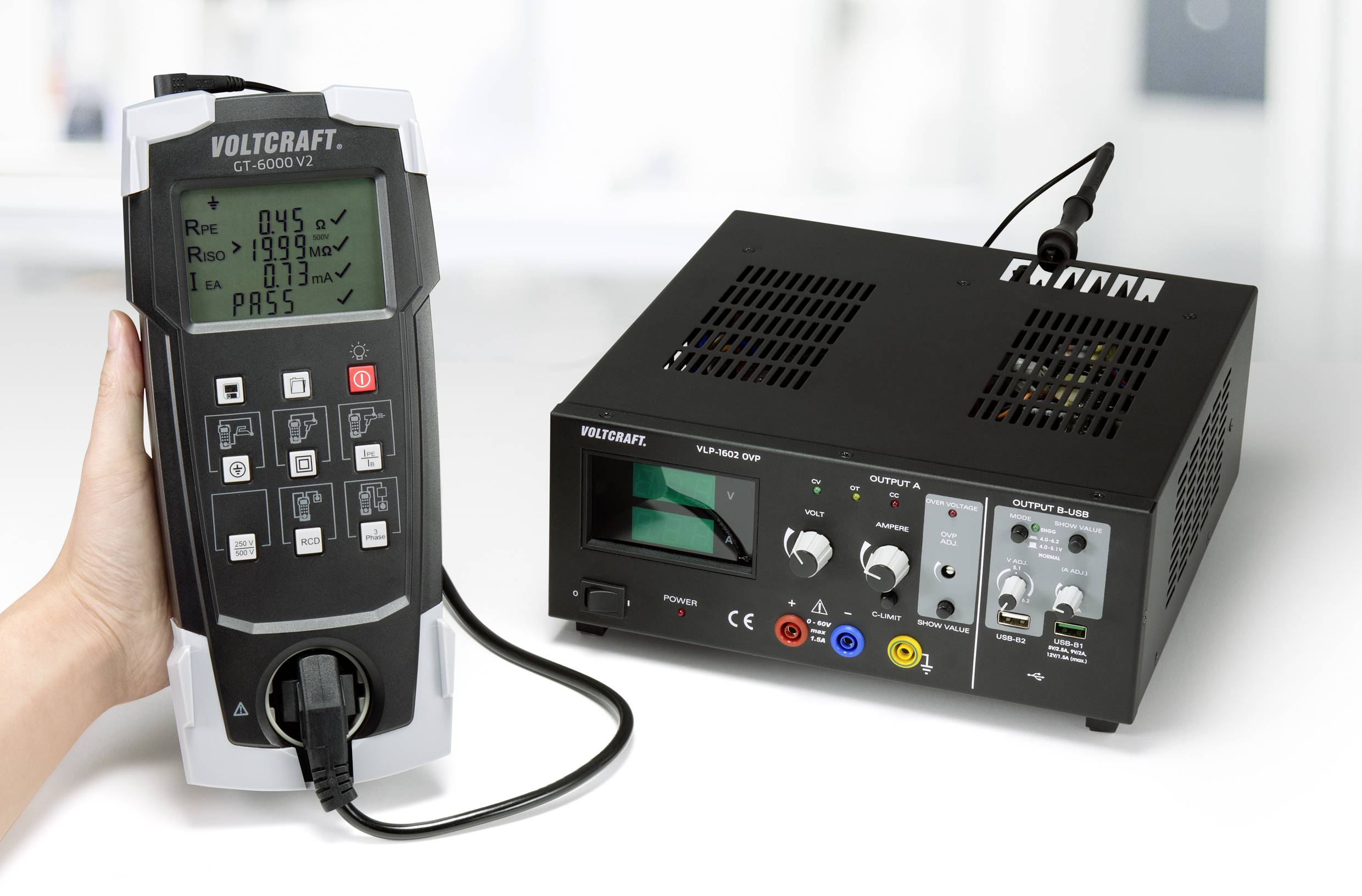 Eine Hand hält ein Prüfgerät neben einem Netzgerät auf einem Tisch. Das Gerät zeigt Testergebnisse auf einem digitalen Display an.