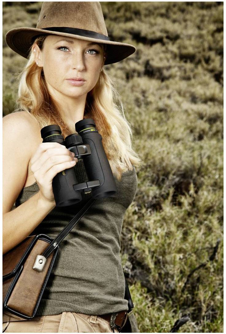 Eine Person mit Hut hält ein Fernglas und steht in einer grasbewachsenen Landschaft.