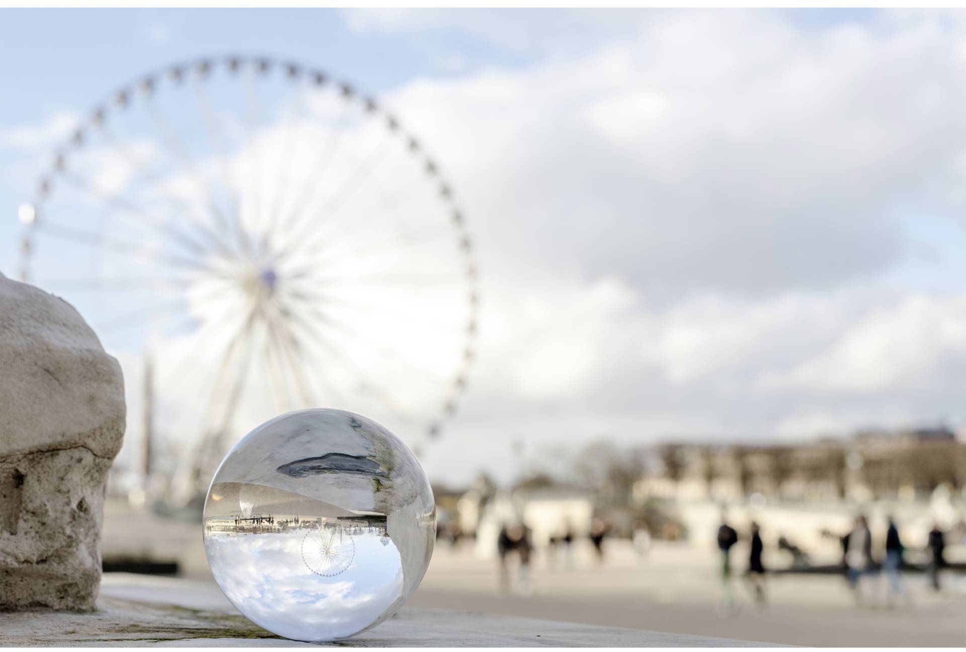 Gläserne Kugel reflektiert den Himmel und Gebäude; im Hintergrund ist ein unscharfes Riesenrad in einer Stadtlandschaft zu sehen.