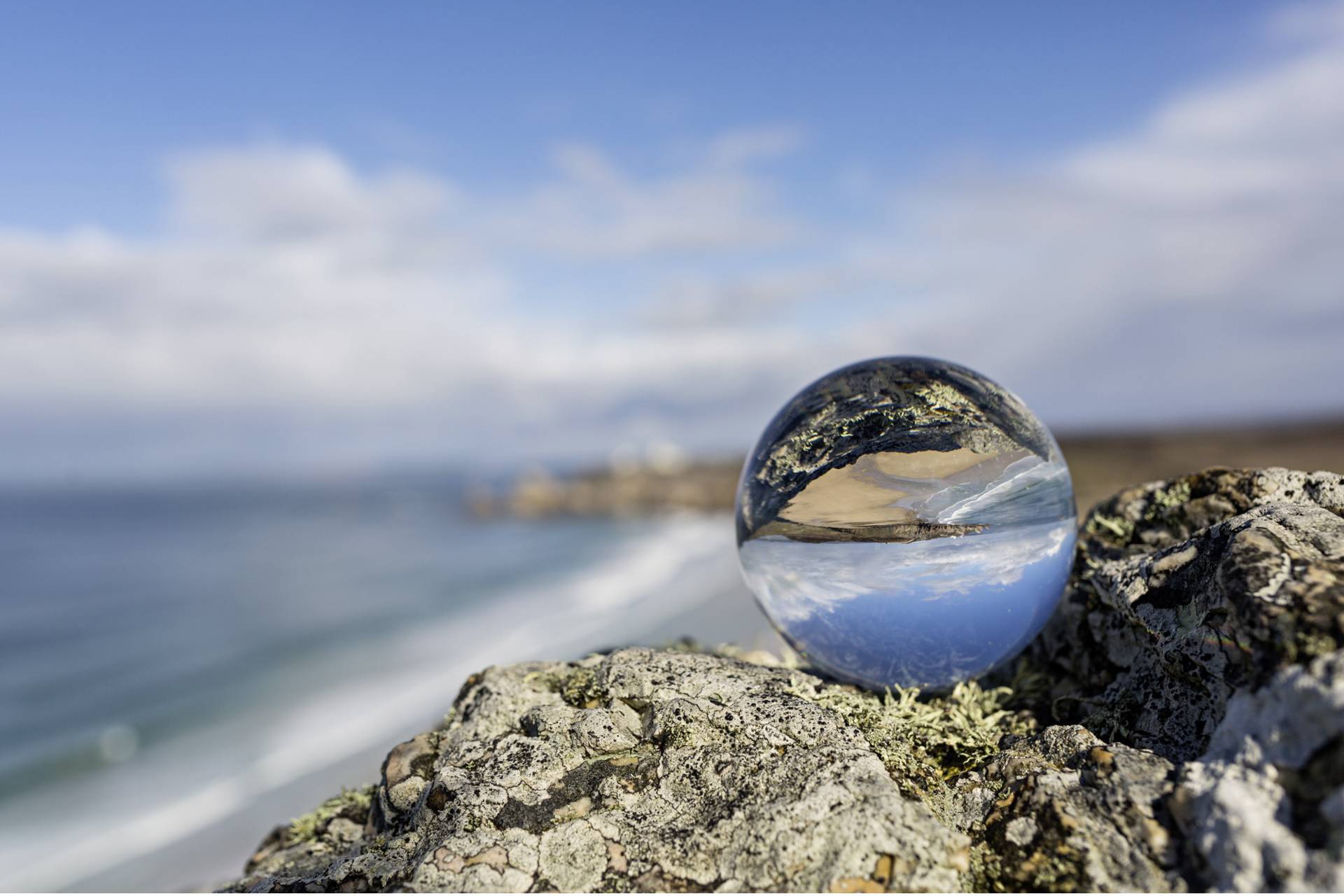 Eine Glaskugel auf einem Felsen spiegelt eine Küstenlandschaft wider, mit Sandstrand und Meer im Hintergrund unter blauem Himmel.