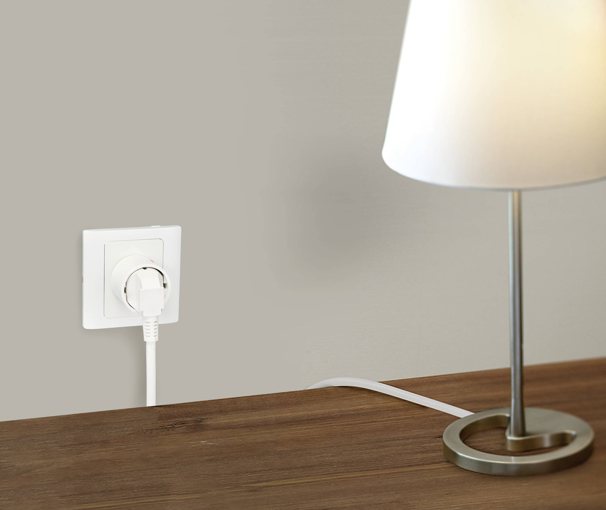 Eine Tischlampe leuchtet auf einem Holztisch. Das Kabel der Lampe ist an eine weiße Wandsteckdose angeschlossen.