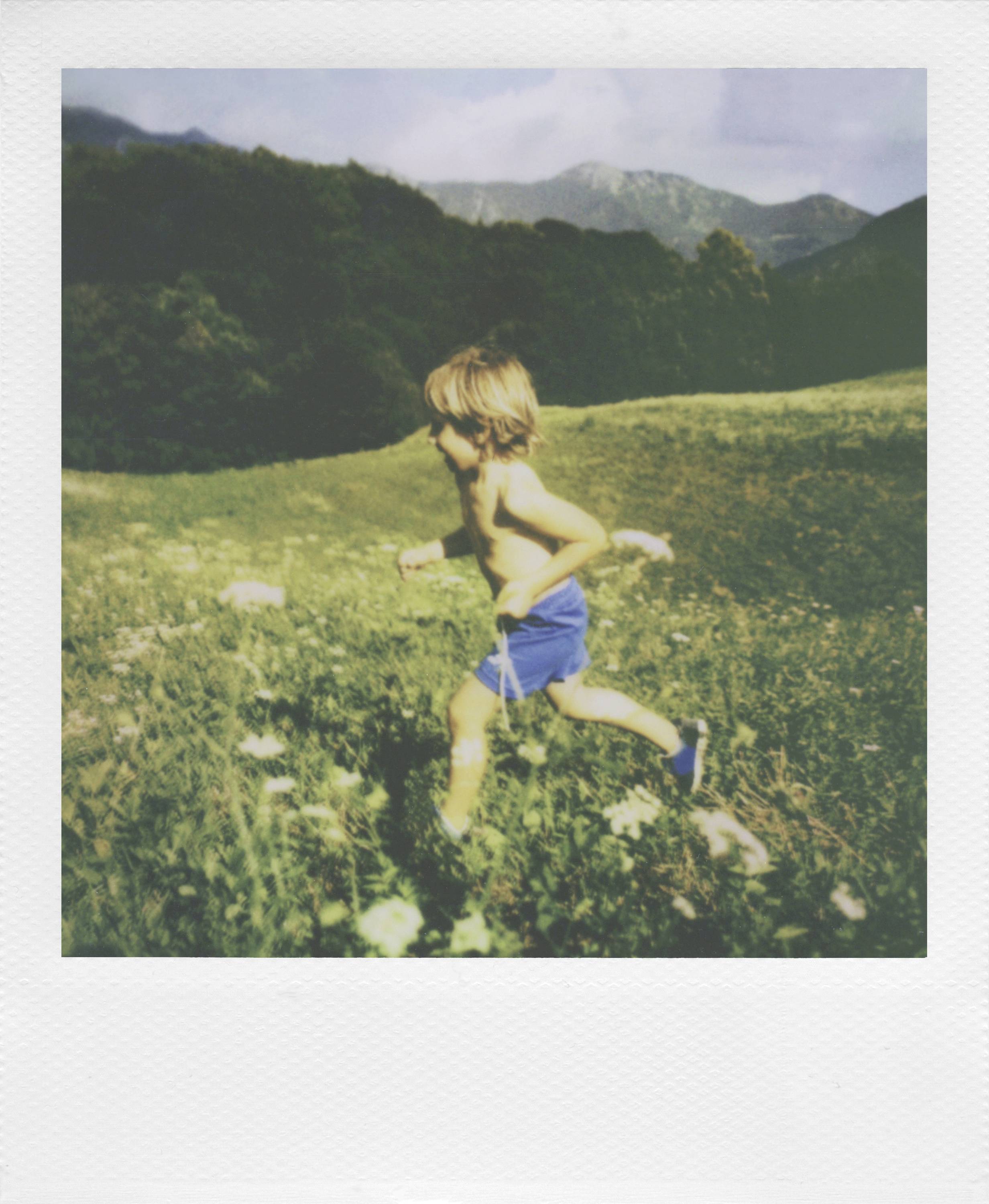 Ein Kind in blauen Shorts läuft barfuß über eine blühende Wiese mit Bergen im Hintergrund. Es wirkt fröhlich und unbeschwert.