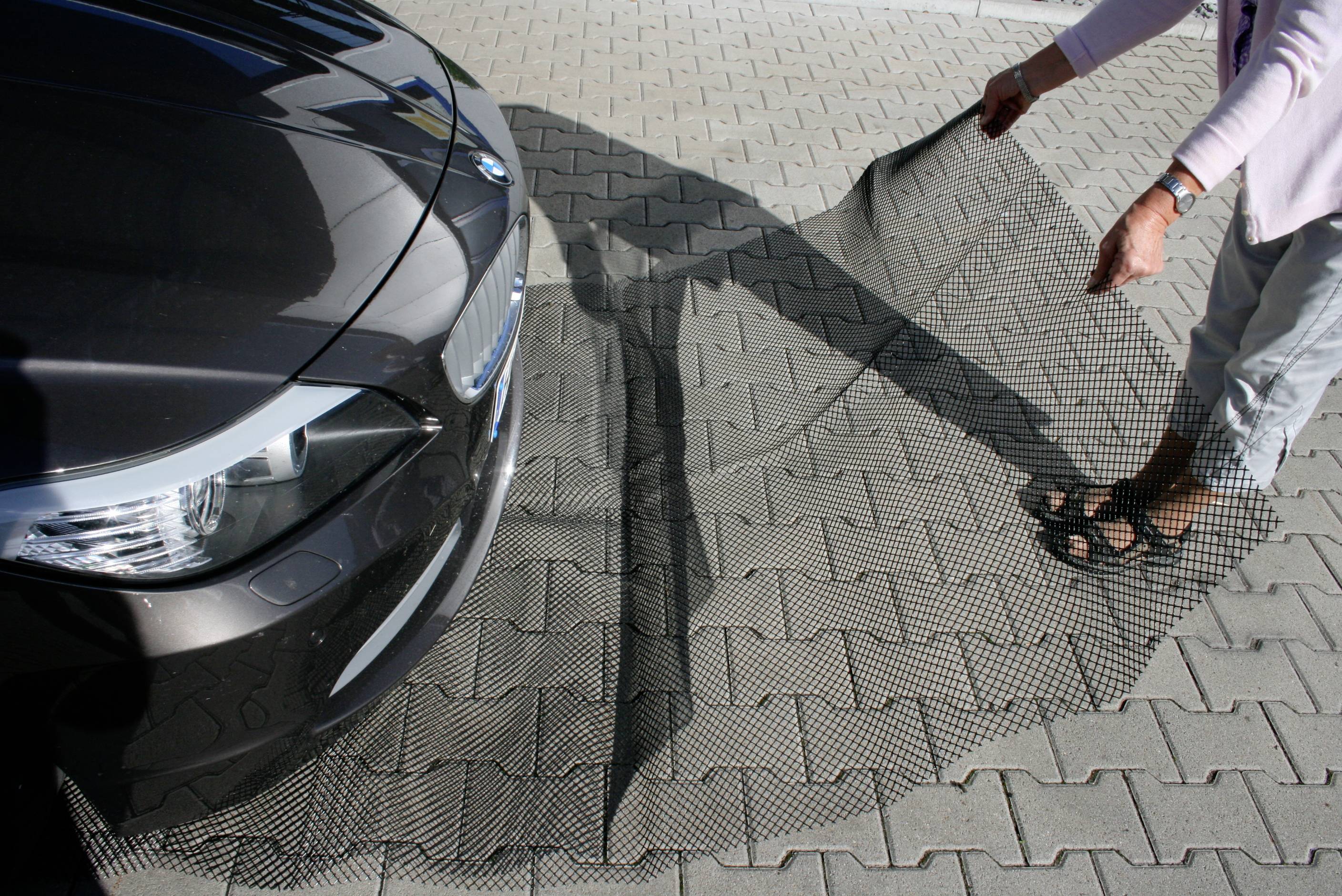 Eine Person legt ein feinmaschiges Netz vor ein geparktes schwarzes Auto auf einem gepflasterten Parkplatz aus.