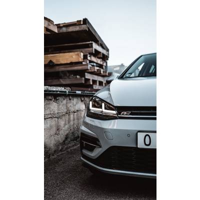 Vorderer Teil eines silbernen Autos neben einem Stapel Holzpaletten. Die Scheinwerfer sind eingeschaltet, an einer Betonwand geparkt.