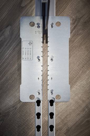 Metallplatte mit Löchern und Skalenmaß, die auf einem Holzboden liegt. Verwendet zur präzisen Markierung oder Montage von Komponenten.