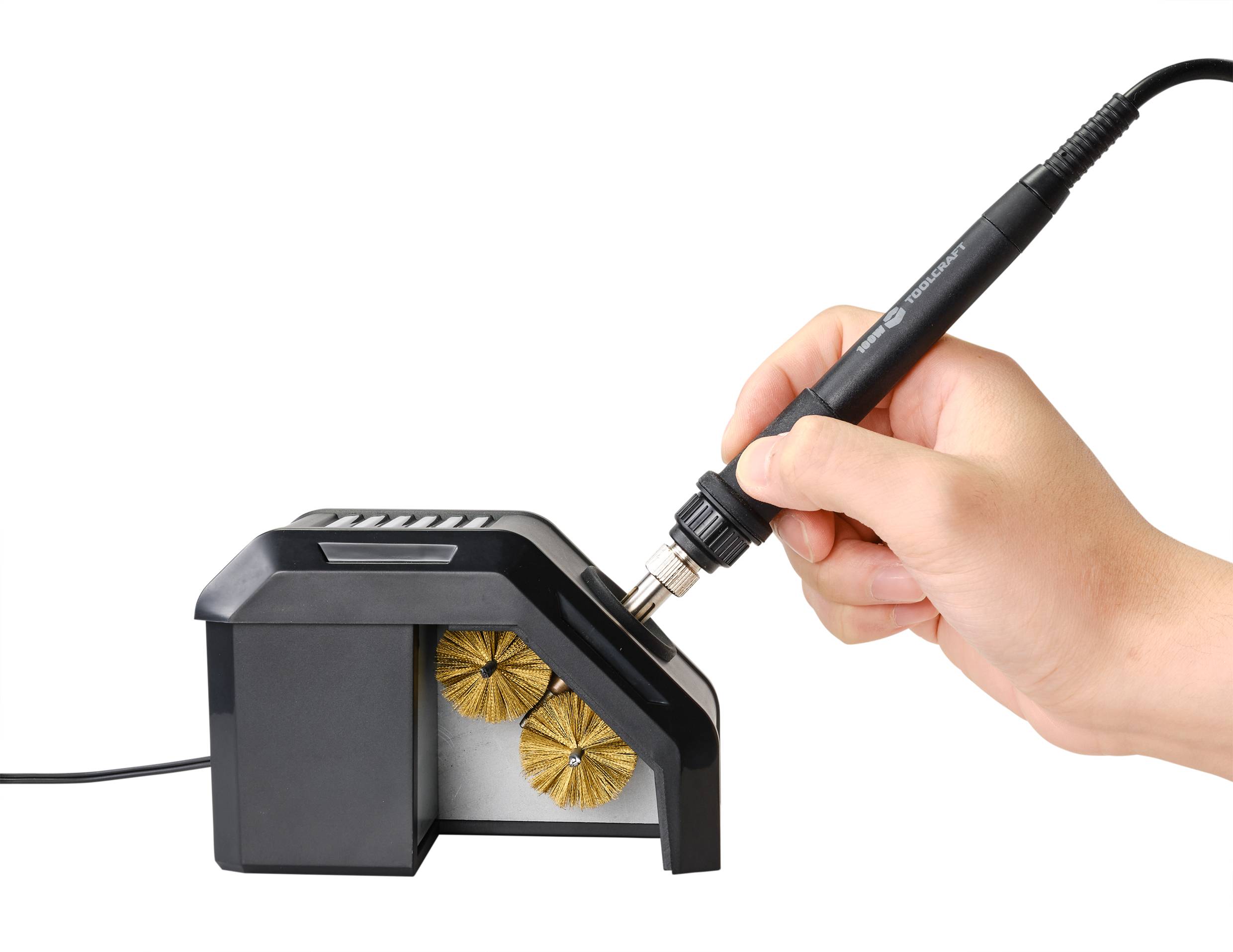 Eine Hand hält einen Lötkolben, der in eine schwarze Lötstation gesteckt wird. Die Station hat zwei goldene Reinigungsbürsten.