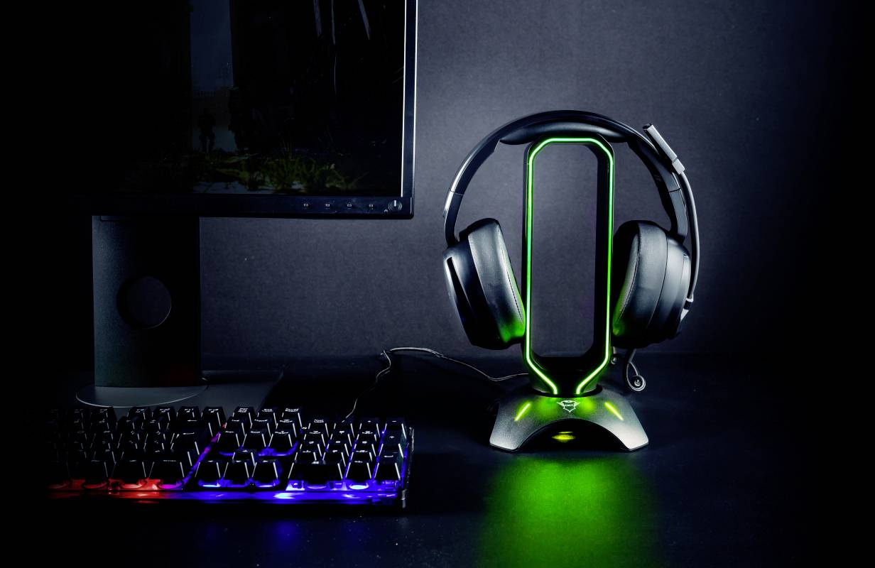Ein beleuchteter Gaming-Kopfhörer steht auf einem Ständer neben einer Tastatur mit farbig beleuchteten Tasten und einem Monitor.