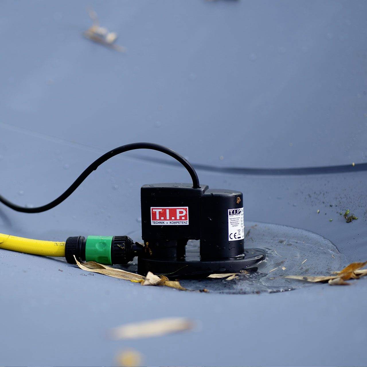 Eine schwarze Wasserpumpe mit rotem 'T.I.P' Logo pumpt Wasser von einer grauen Oberfläche; daran angeschlossen ein gelber Schlauch.