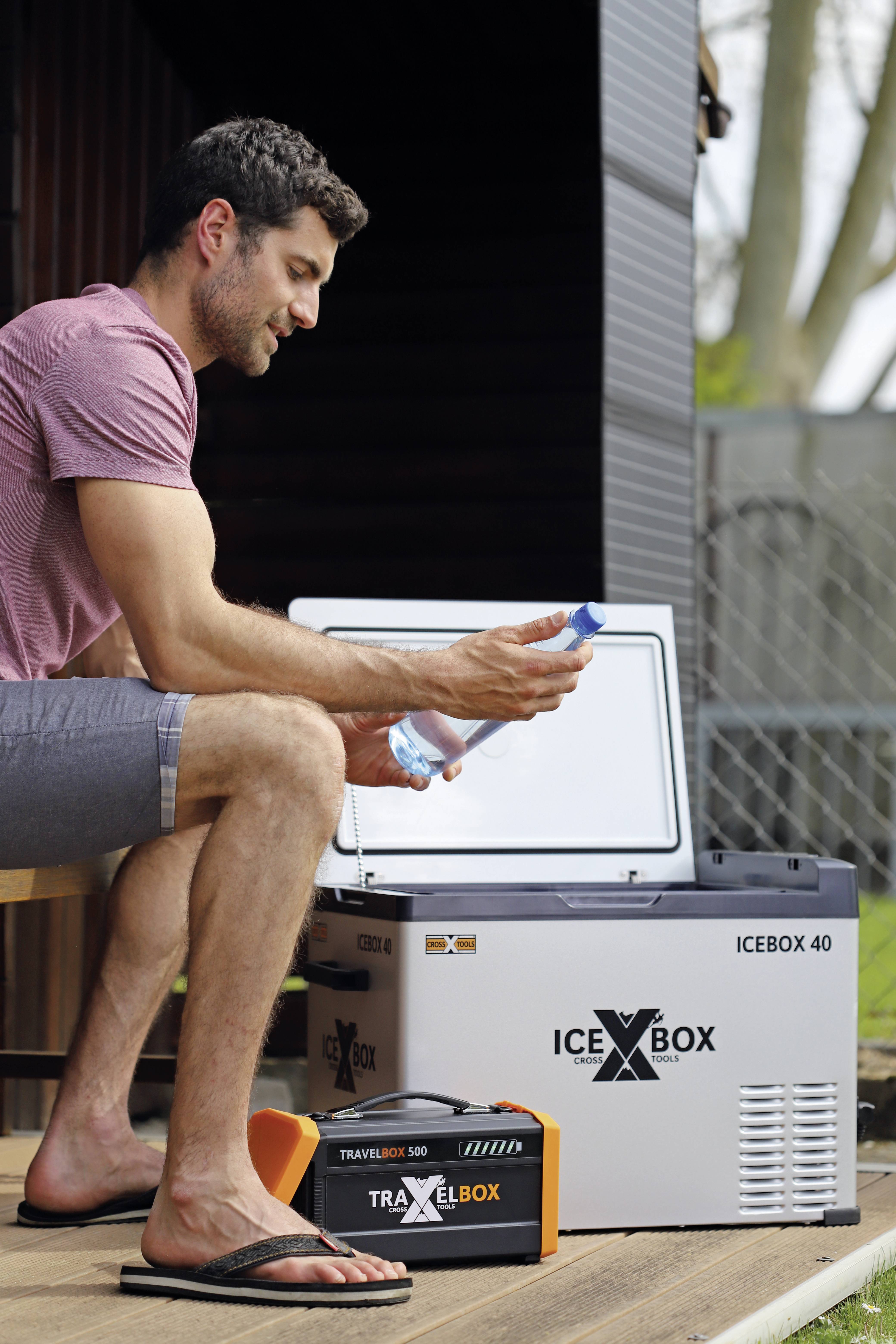 Ein Mann sitzt neben einer geöffneten Kühlbox und hält eine Wasserflasche in der Hand. Im Hintergrund ist ein Garten zu sehen.