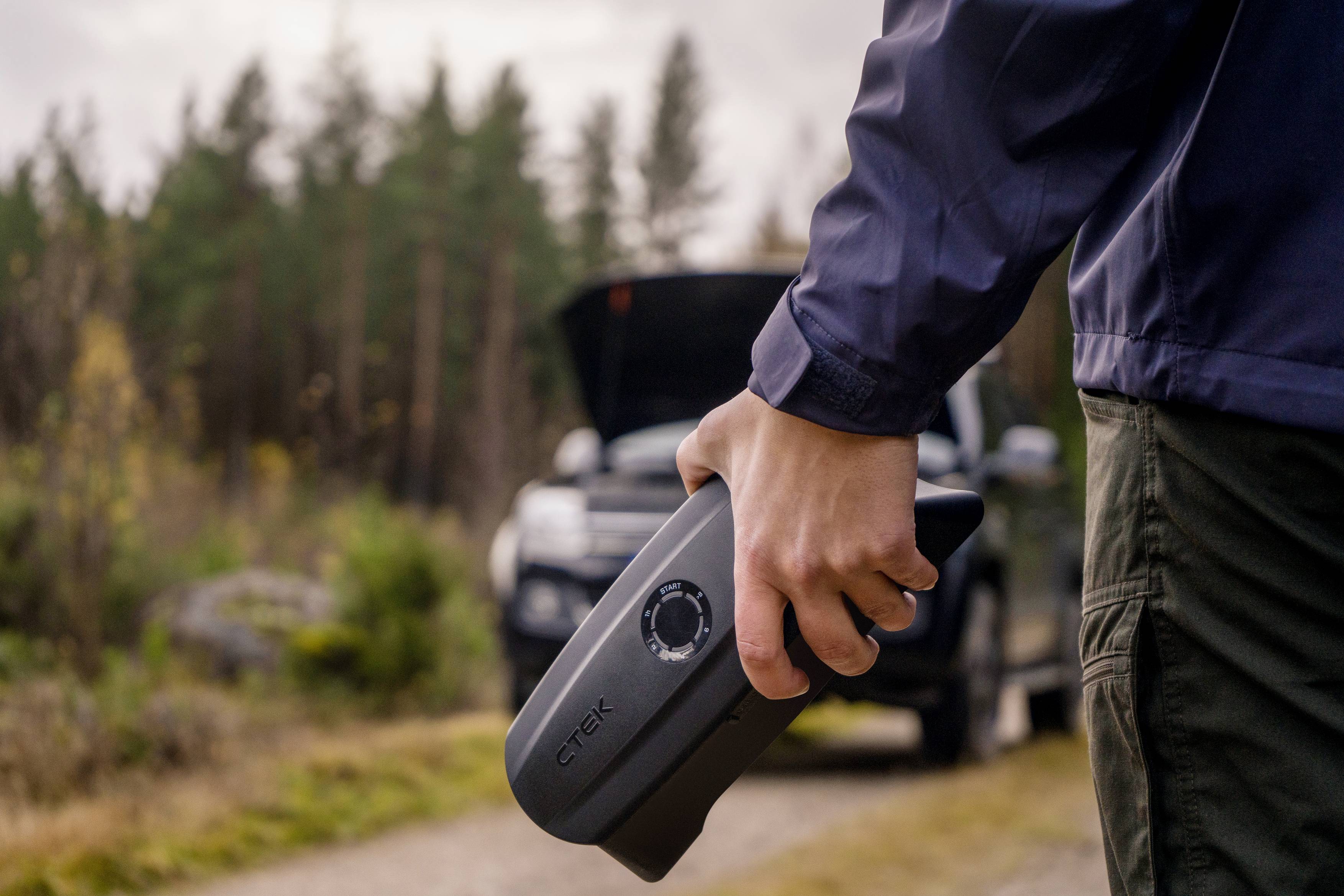 Person hält ein Batterieladegerät, während ein Auto mit geöffneter Motorhaube im Hintergrund zu sehen ist. Wälder umgeben die Szene.
