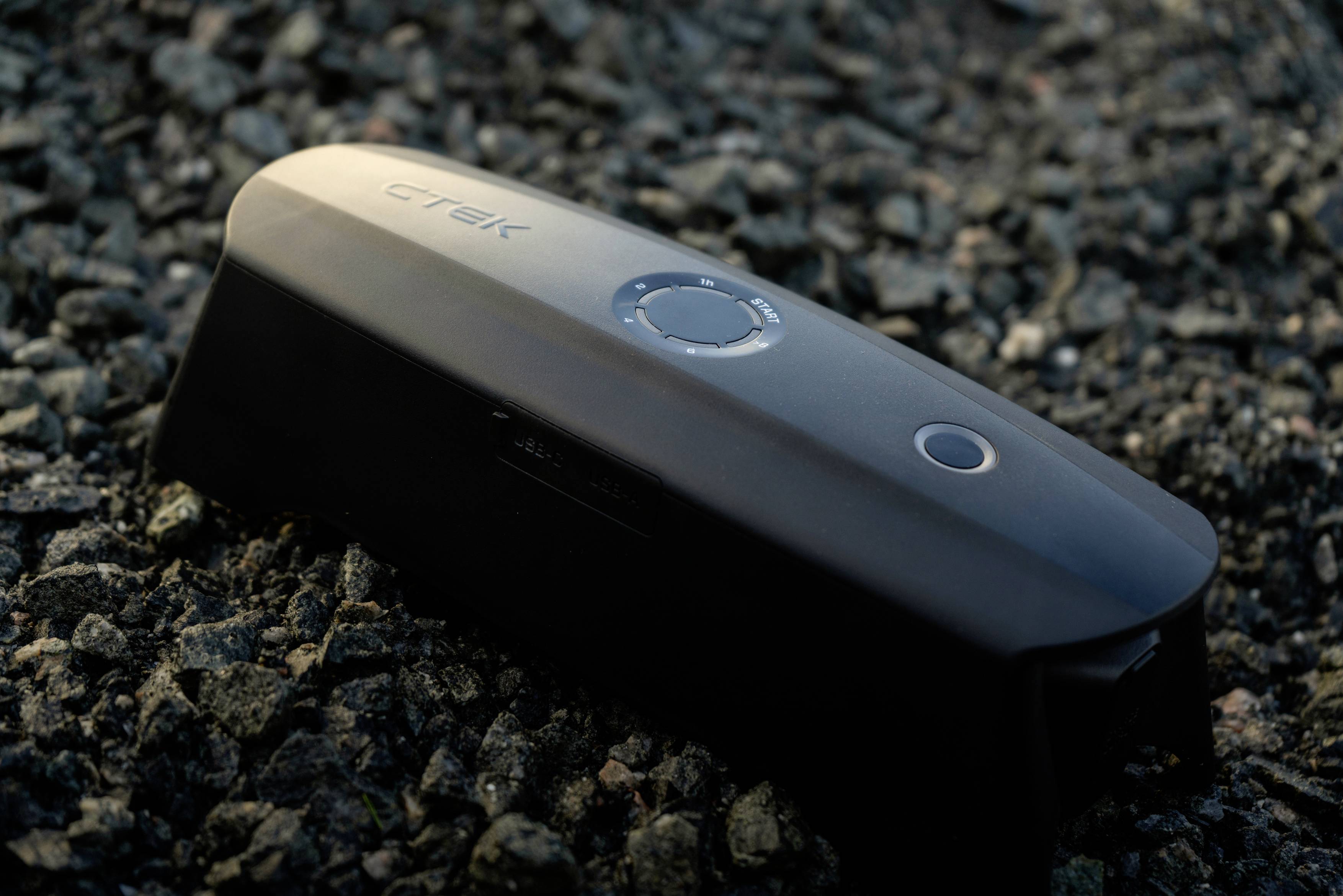 Ein schwarzes technisches Gerät liegt auf Kies. Es hat eine runde Taste und das Logo 'CTEK'. Es könnte ein Ladegerät sein.
