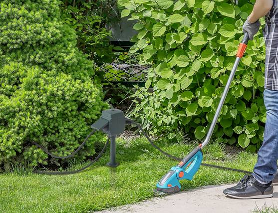 Gärtner arbeitet mit einem elektrischen Trimmer, der über eine wetterfeste Gartensteckdose im Grünen mit Strom versorgt wird.