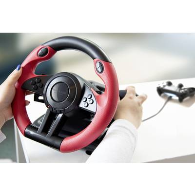 Person hält ein rotes Gaming-Lenkrad mit Tasten, eine Spielekonsole und Controller sind im Hintergrund zu sehen.