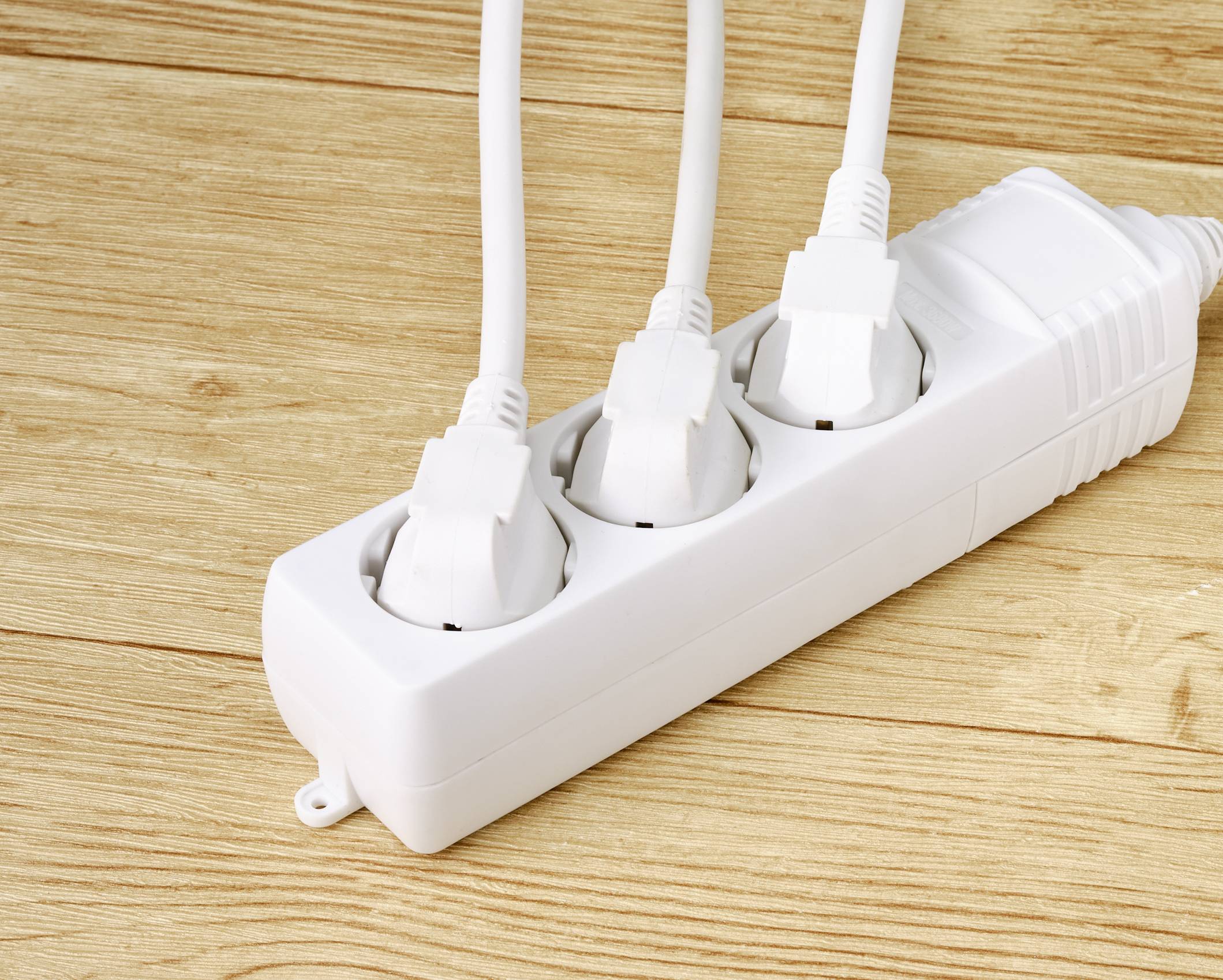 'Drei weiße Stecker sind in eine weiße Mehrfachsteckdose auf einem Holzboden eingesteckt.'