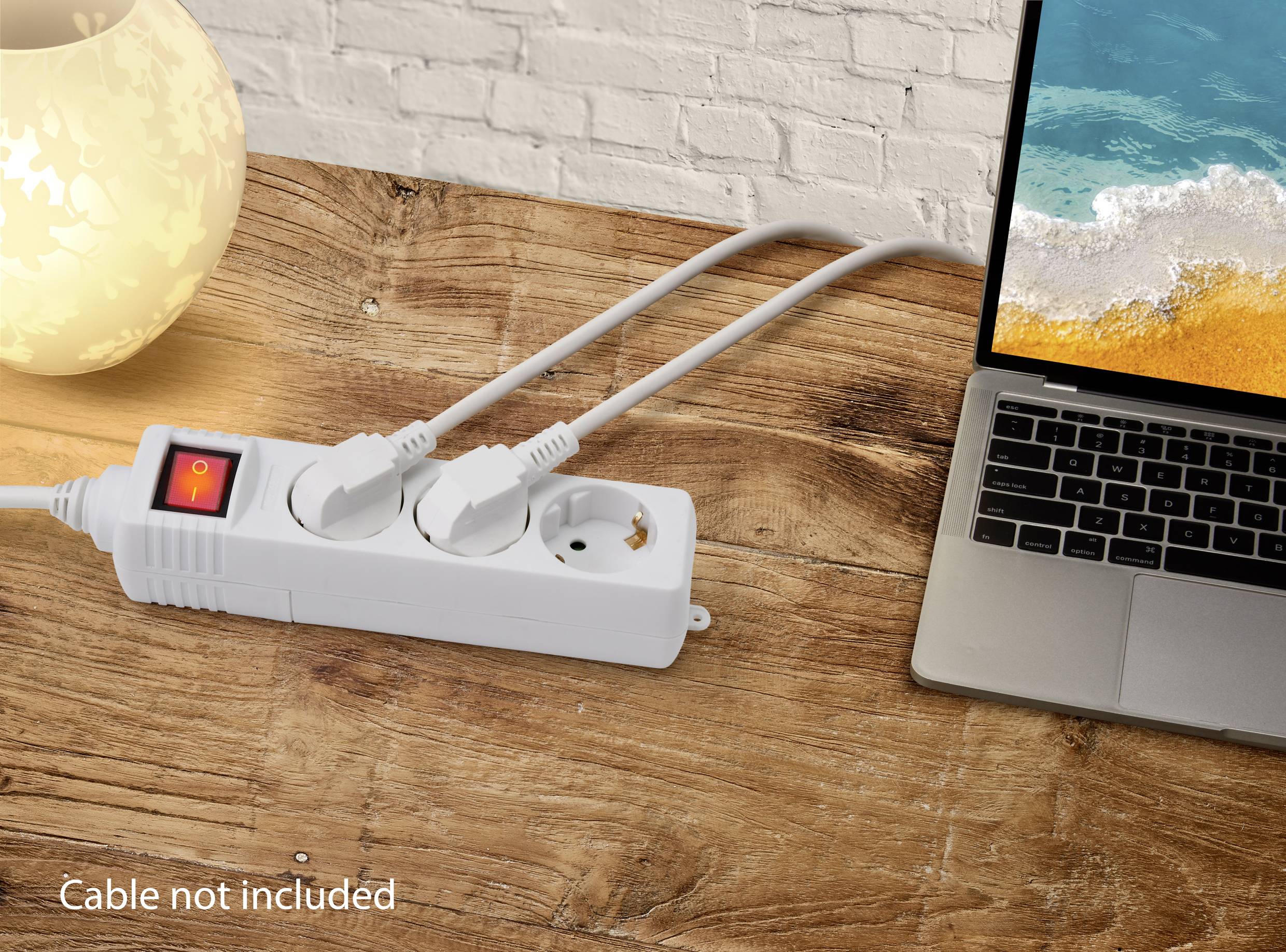 Eine weiße Mehrfachsteckdose mit zwei eingesteckten Kabeln liegt auf einem Holztisch neben einem eingeschalteten Laptop und einer Lampe.