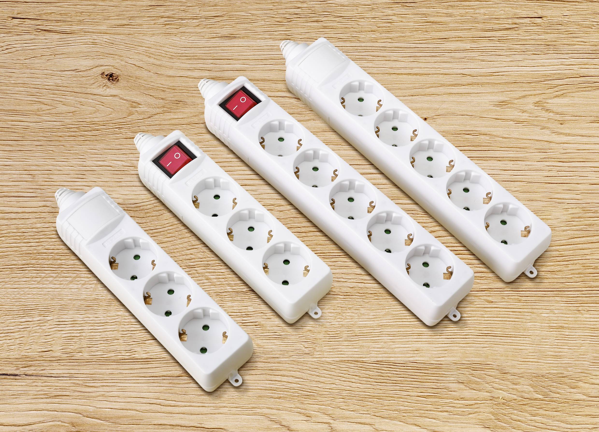 Vier weiße Mehrfachsteckdosen mit Schaltern liegen auf einem Holztisch. Jede Steckdosenleiste hat zwischen drei und sechs Steckplätze.