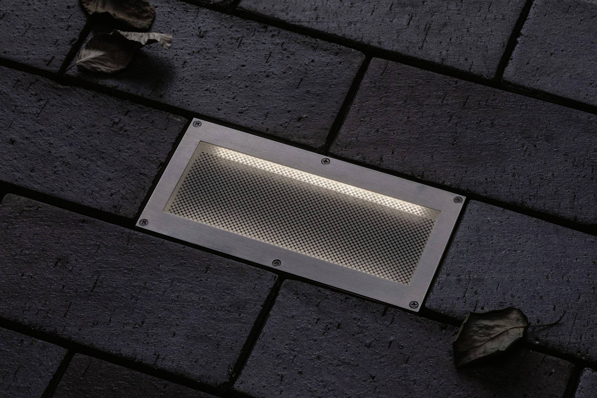 Rechteckige Bodenlampe in dunklem Pflastersteinbelag, beleuchtet, mit zwei trockenen Blättern in der Nähe.