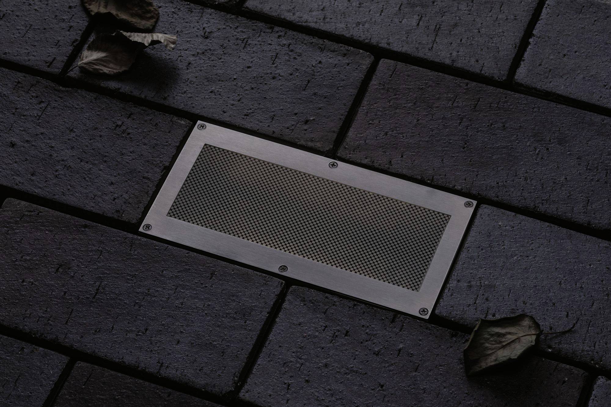 Eine rechteckige Metallabdeckung in einem dunklen Pflastersteinweg, umgeben von zwei trockenen Blättern.
