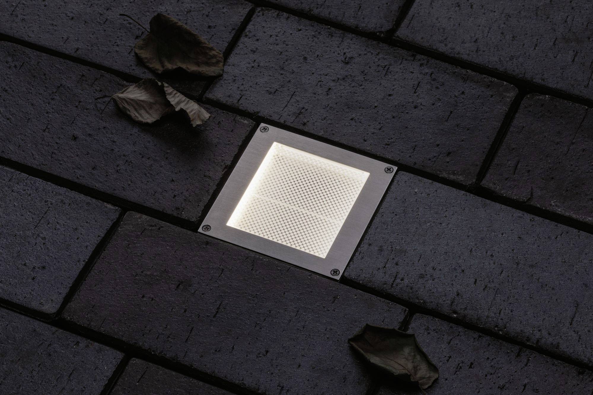 Beleuchtetes quadratisches Pflastersteinlicht in dunklem, gepflastertem Gehweg, umgeben von Blättern.