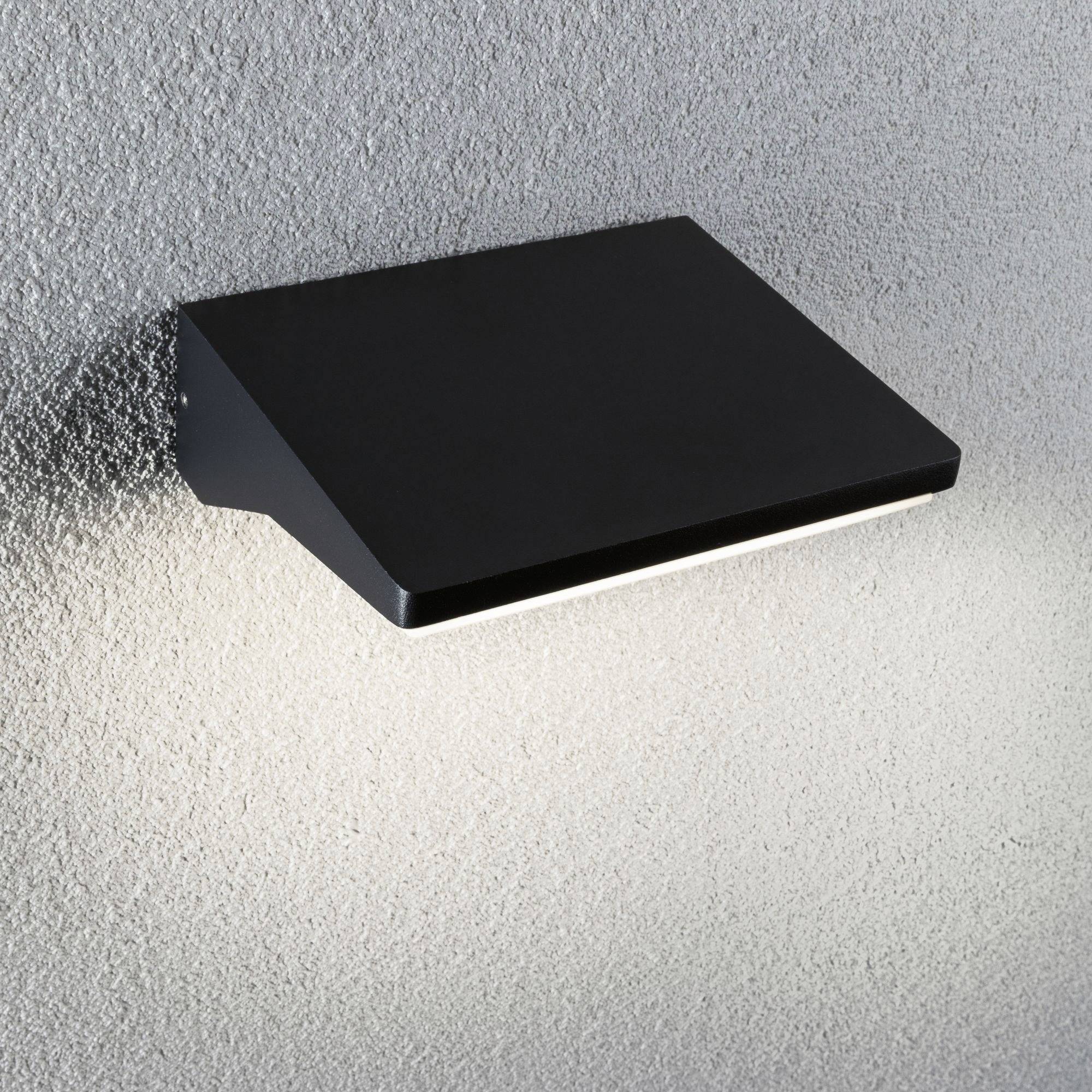 Eine schwarze, rechteckige Wandleuchte auf einer grauen Oberfläche leuchtet nach unten und erzeugt einen Lichtschein.