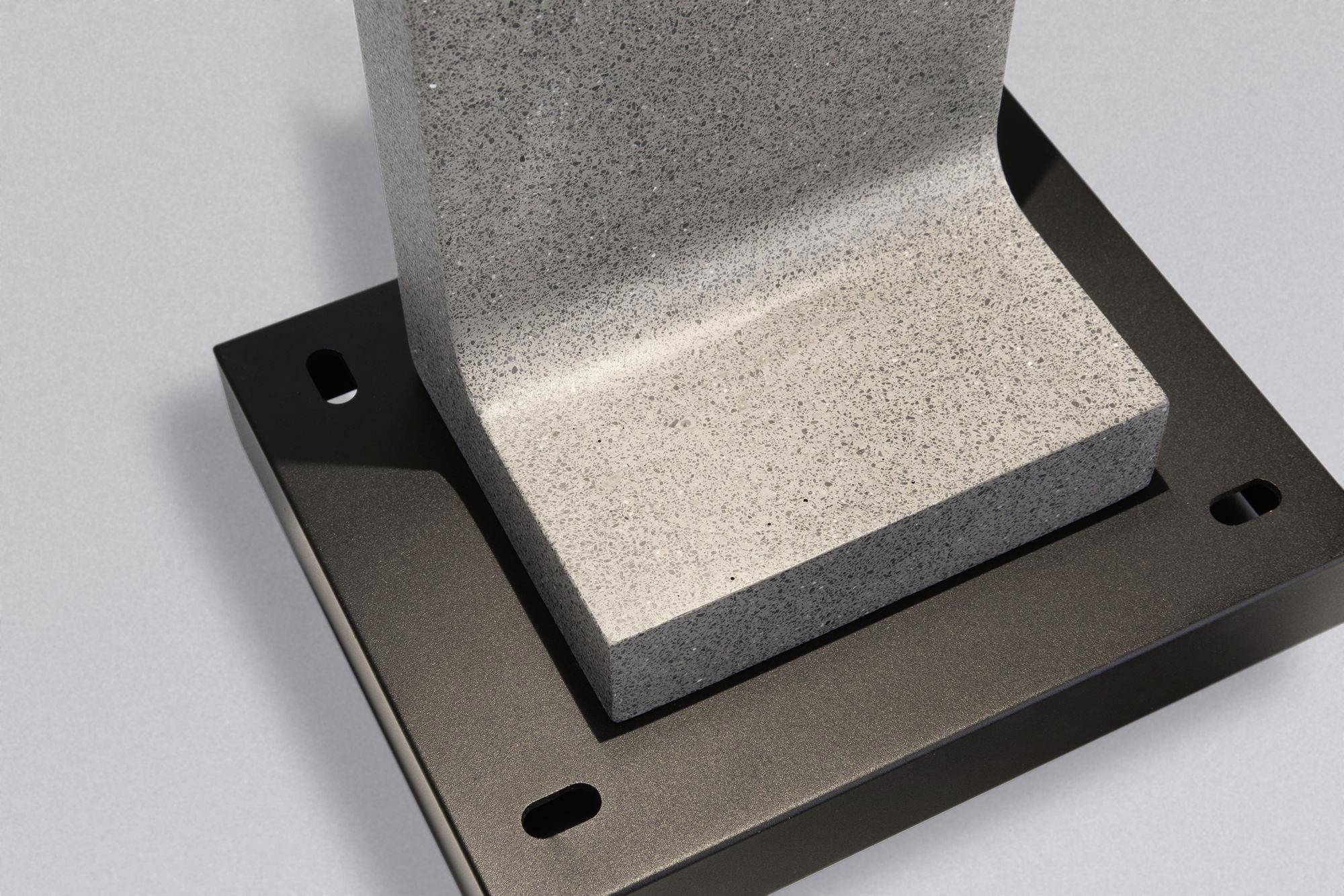 Sockel aus grauem Beton auf einer schwarzen Metallbasis mit vier Löchern, wahrscheinlich zur Befestigung an einer Oberfläche.