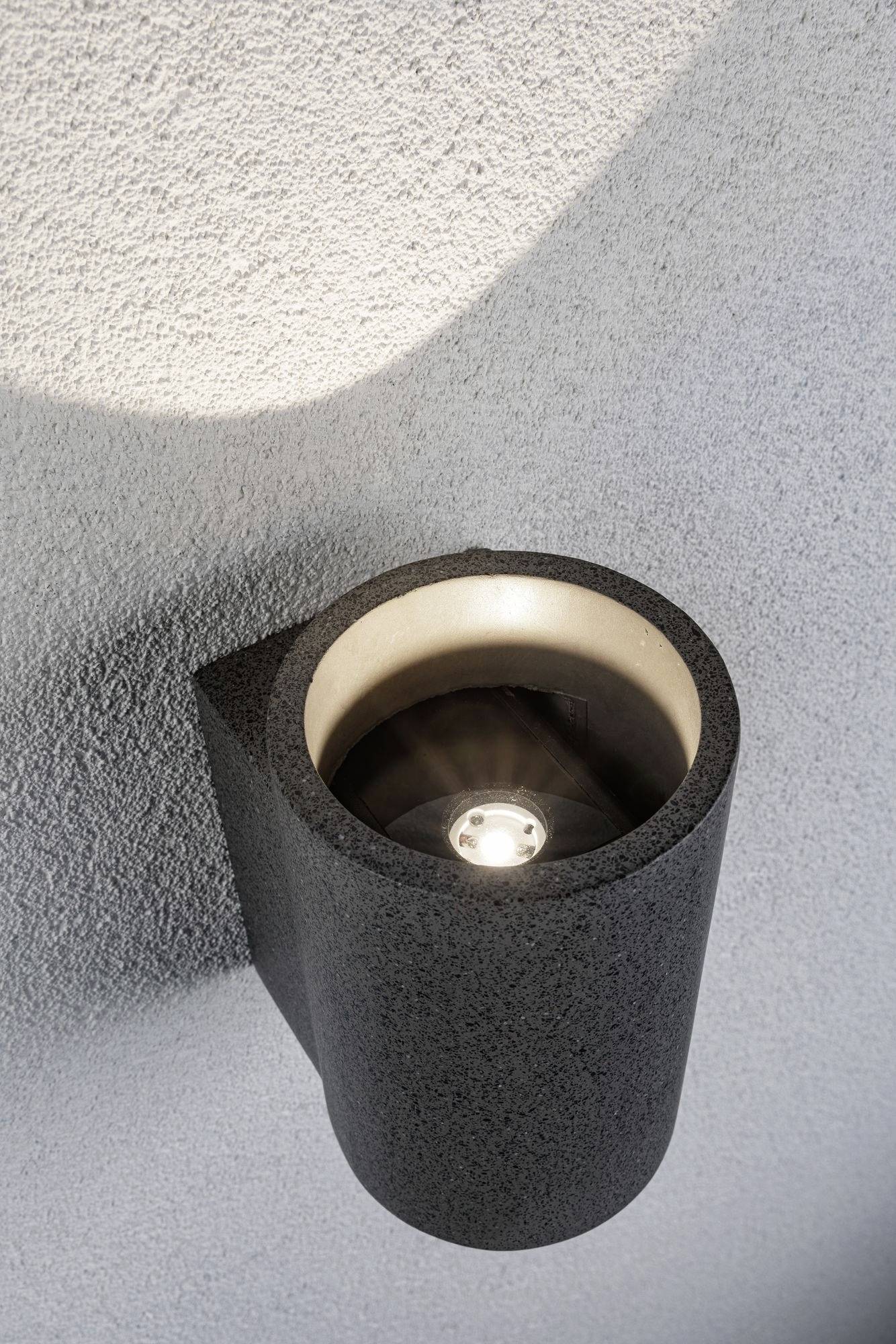 Moderne, zylindrische Außenlampe mit schwarzem Gehäuse an einer grauen Wand montiert, strahlt nach oben gerichtetes Licht aus.