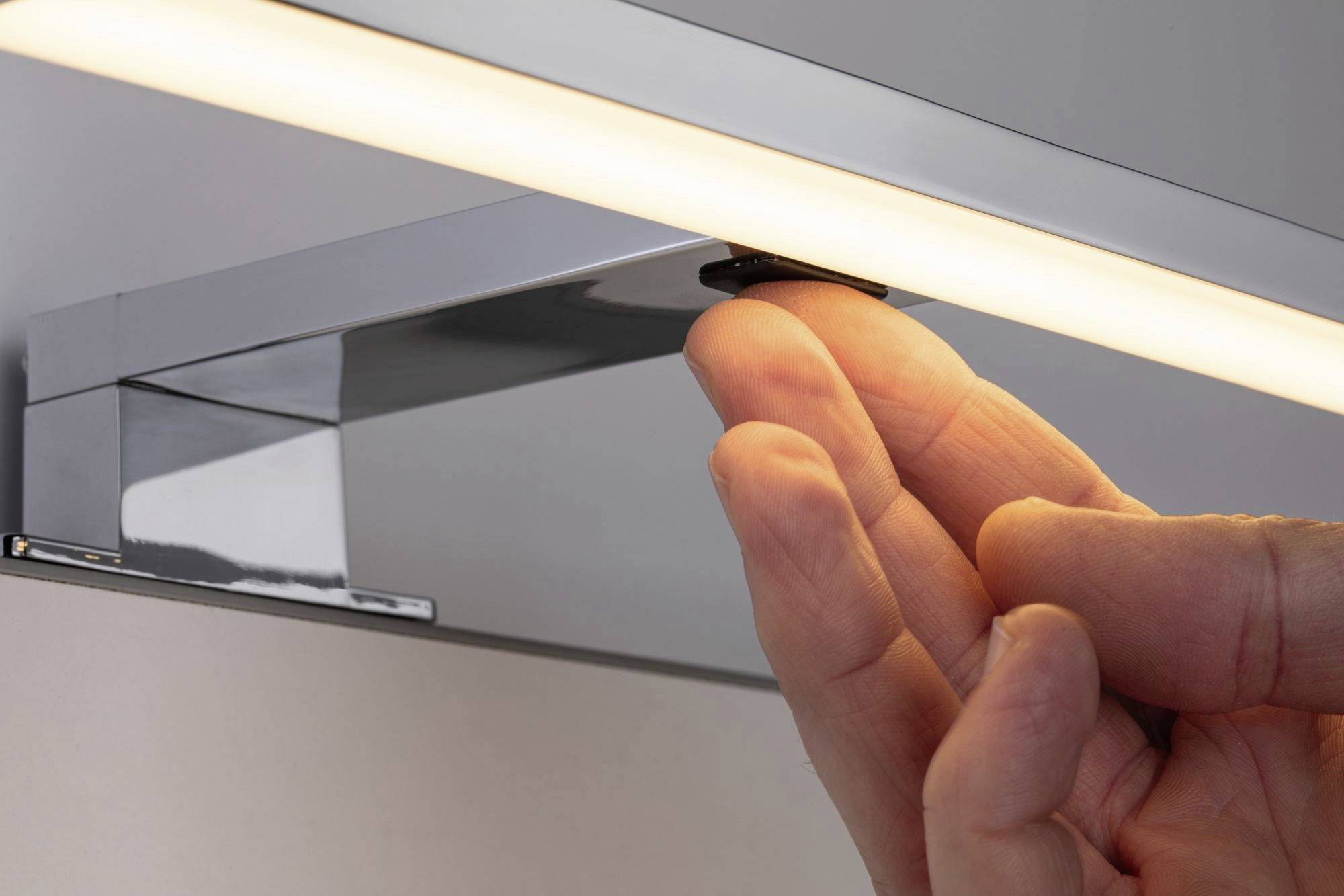 Eine Hand drückt den Schalter einer modernen, rechteckigen LED-Leuchte an der Wand. Die Lampe strahlt warmes Licht aus.