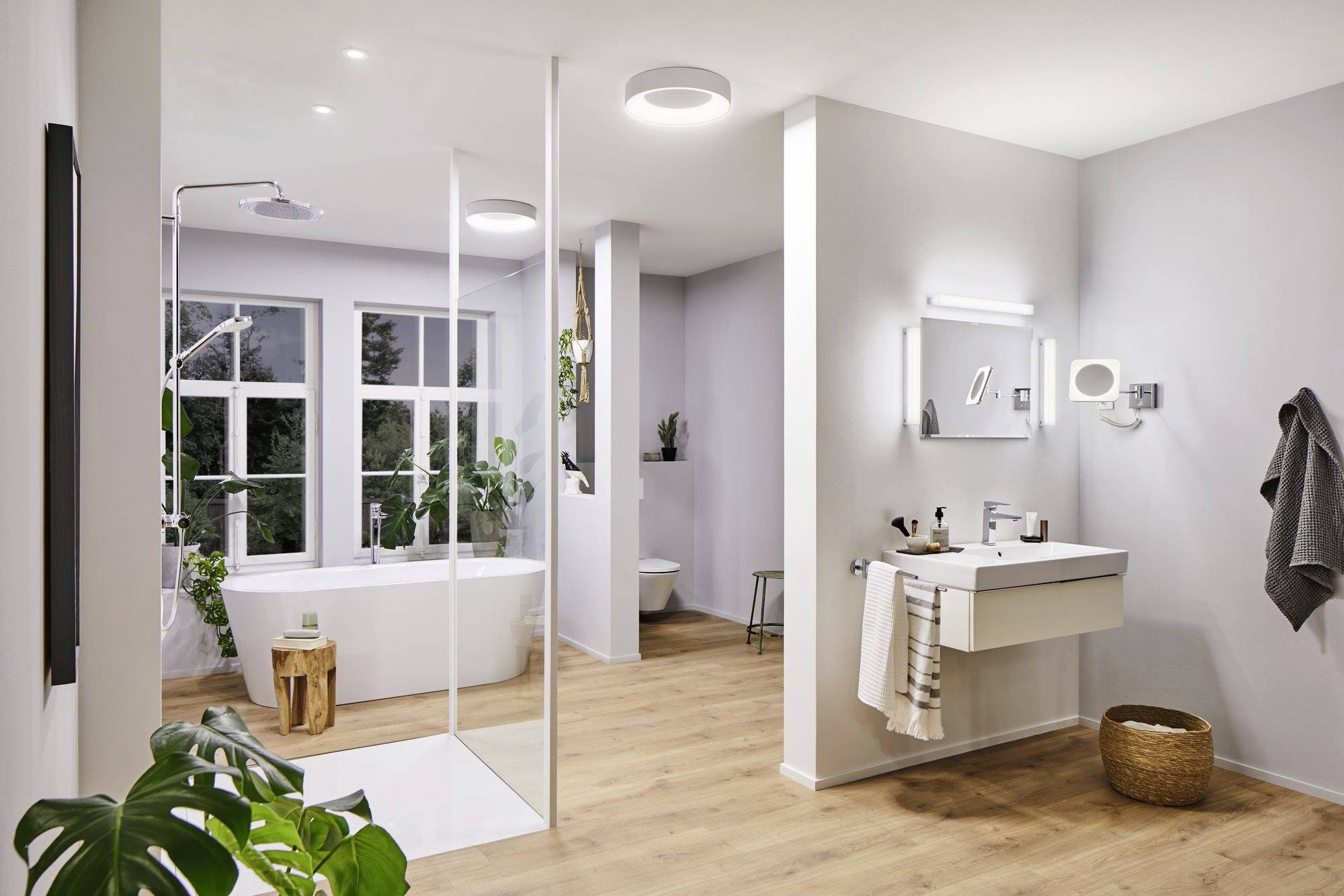 Modernes Badezimmer mit freistehender Badewanne, großer Dusche, Spiegel über Waschbecken und grünen Pflanzen. Helle, minimalistische Einrichtung.